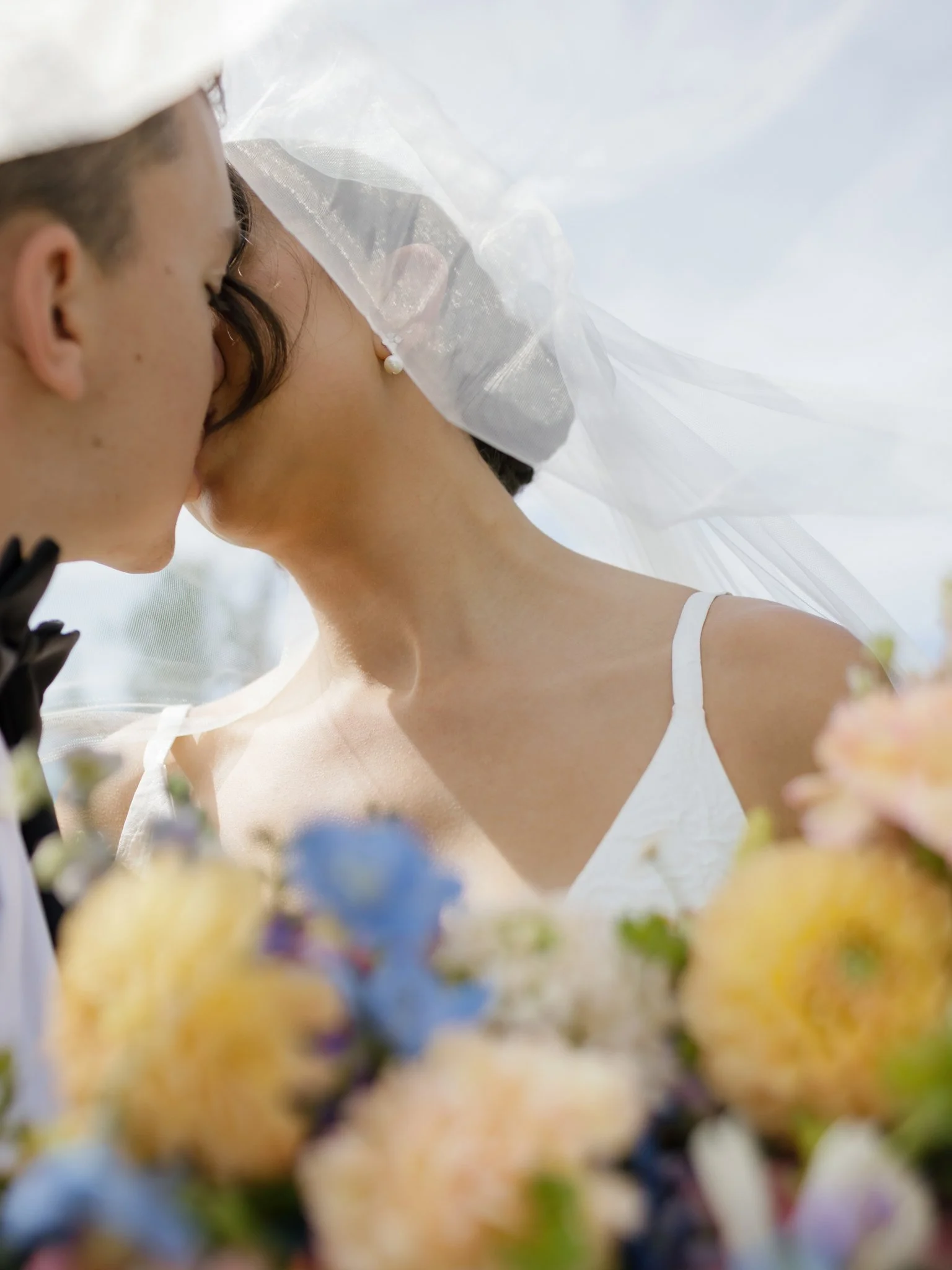 A bride and groom kissing outdoors, with a veil and wedding dress visible, and flowers in the foreground in CDA, Idaho.