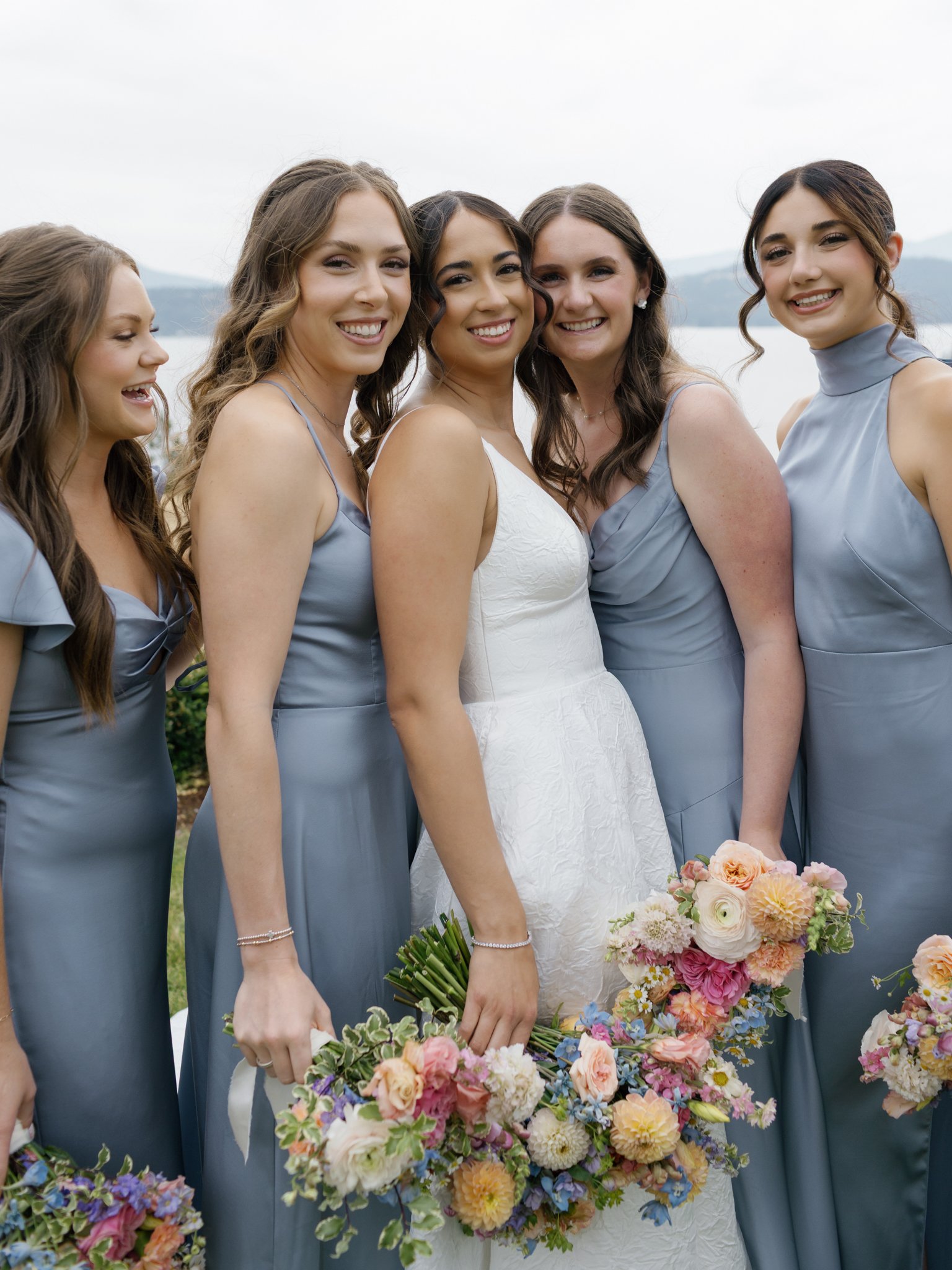 A group of five women, four bridesmaids in blue dresses and one bride in white, standing outdoors near water and holding bouquets of colorful flowers, smiling at the camera at Elk Point wedding above Lake Coeur d’Alene.