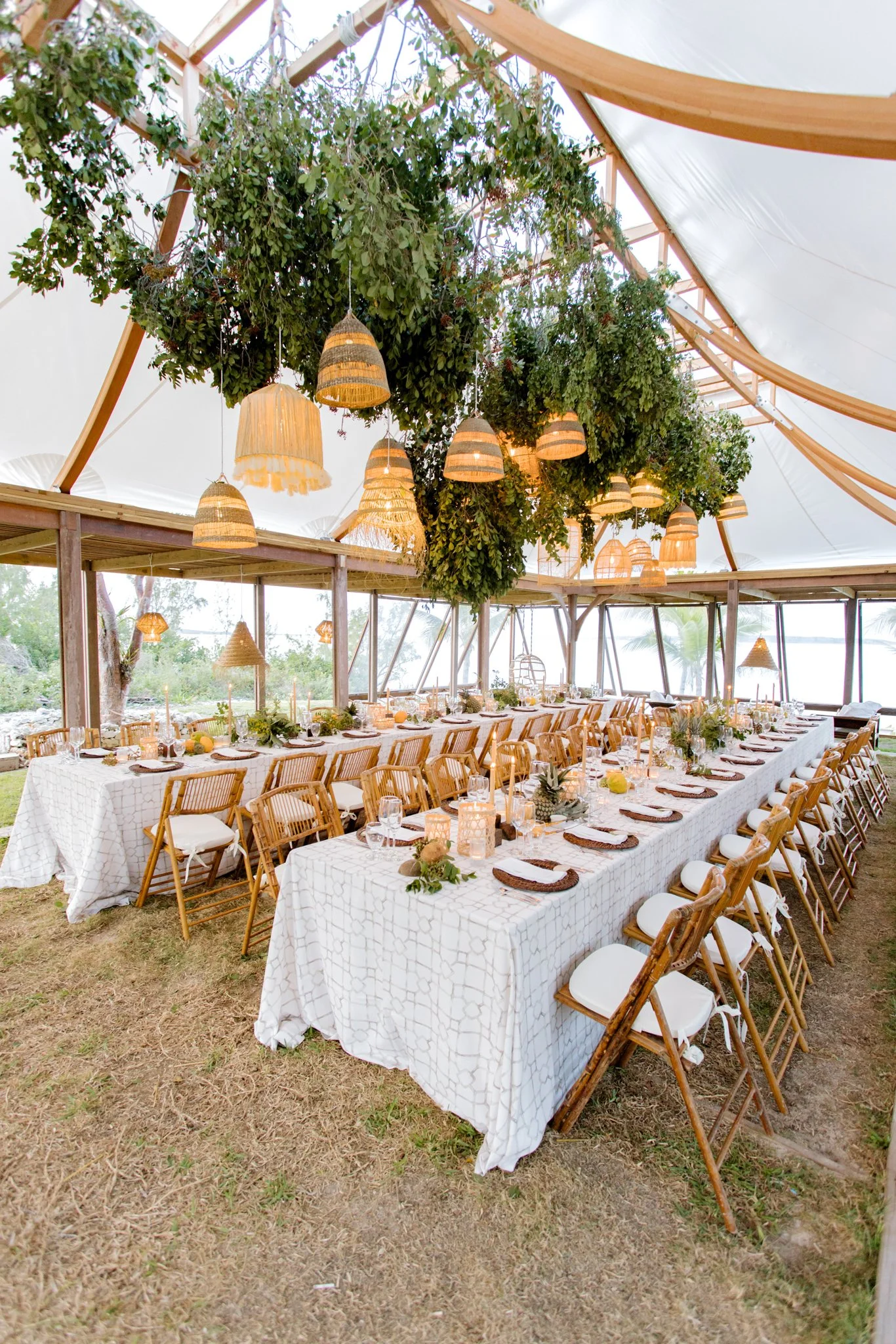 Indoor event space decorated for a feast, with long tables covered in white tablecloths, wooden chairs, candle lanterns, and hanging woven pendant lights, surrounded by greenery overhead.