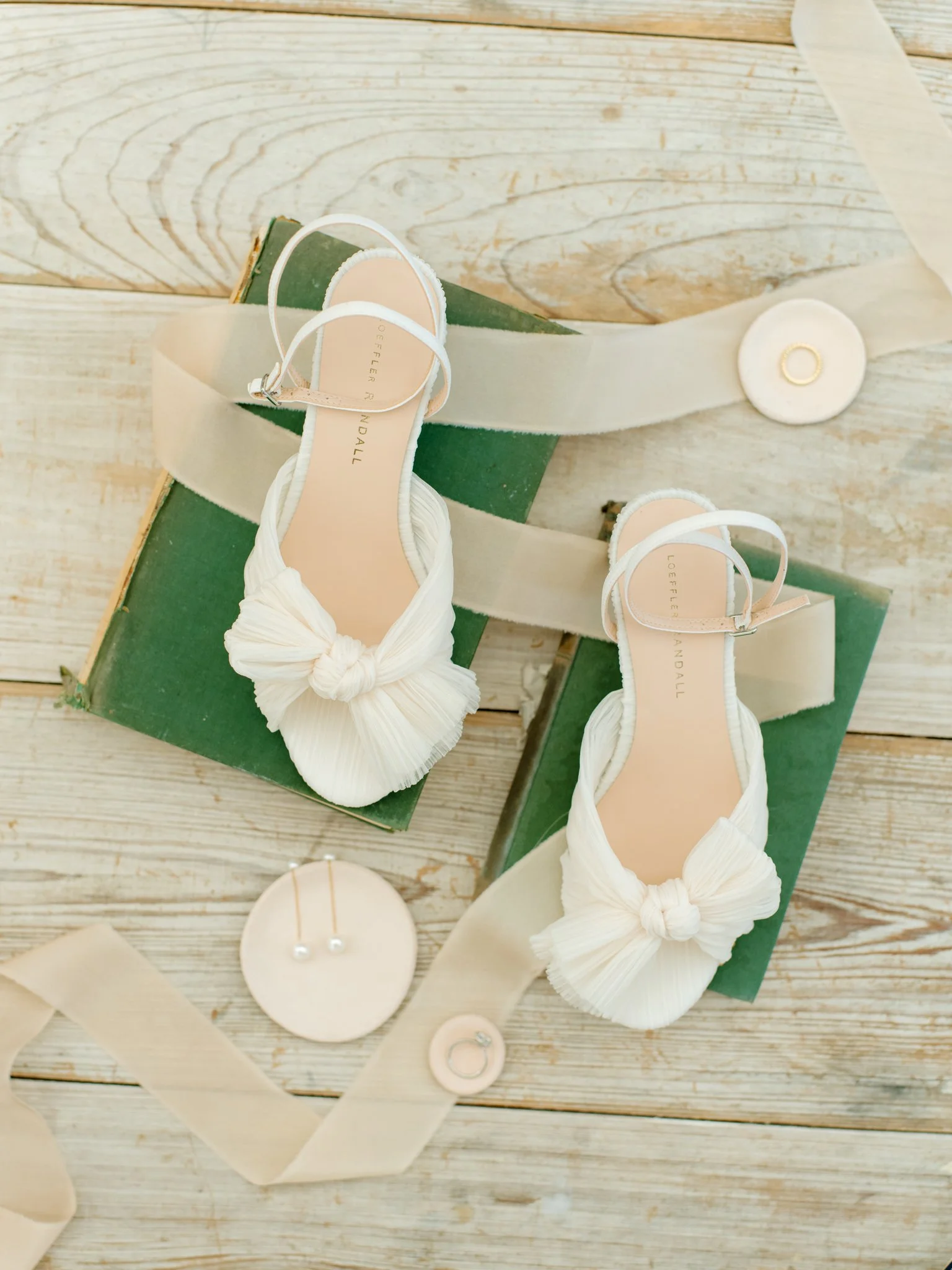 A pair of white ruffled bow sandals on green boxes, surrounded by beige ribbons, pearl earrings, and a pearl headband, all on a light wooden surface  at The Other Side Hotel Eleuthera during a luxury Bahamas destination wedding.