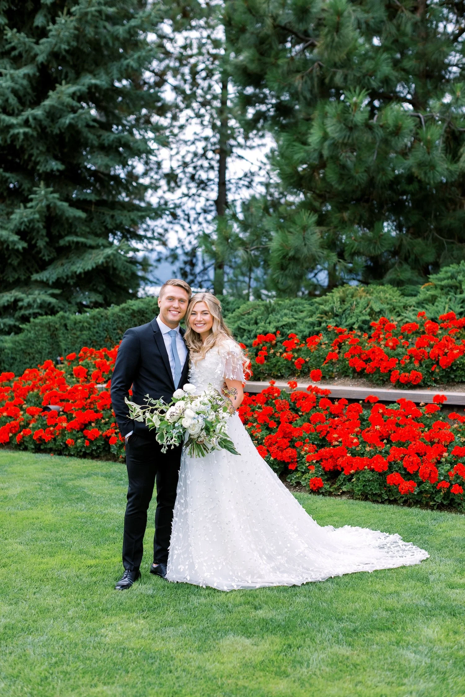 A bride and groom dressed in wedding attire, standing on a lush green lawn with vibrant red flowers and tall trees in the background, smiling at the camera.
