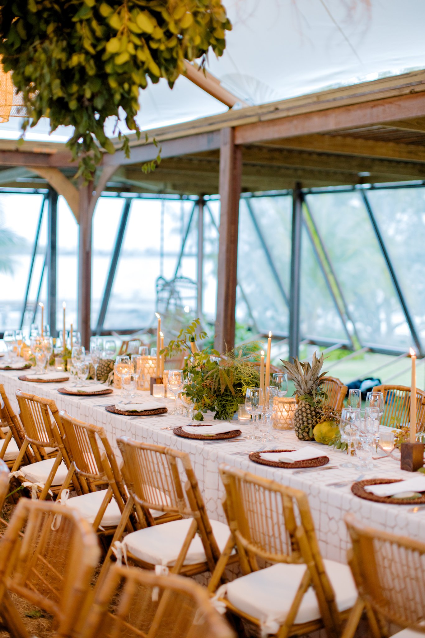 A decorated outdoor dining table with candles and fruit, set inside a greenhouse-like structure with wooden beams and transparent panels.