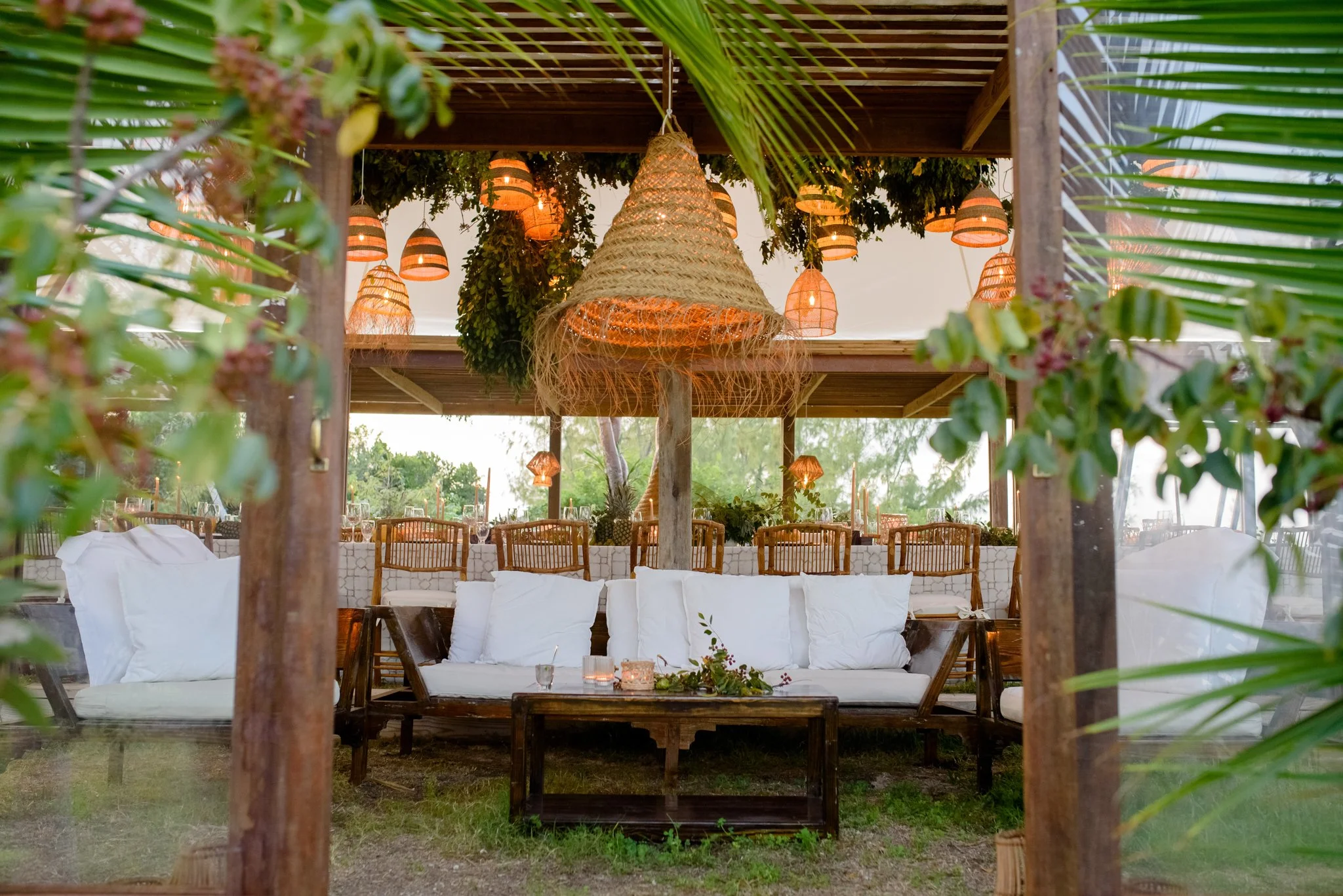 Outdoor dining area with wooden furniture, white cushions, hanging woven lanterns, and lush greenery during daytime.