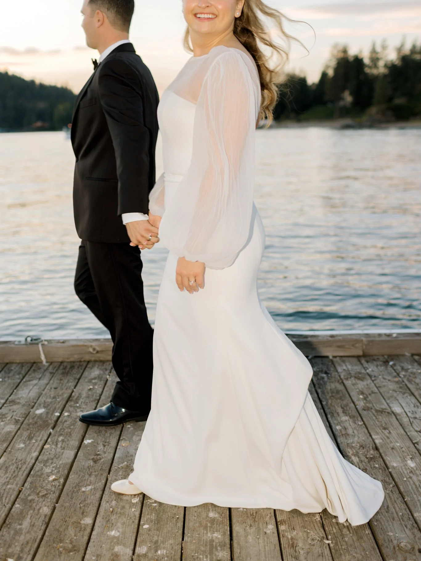 A bride in a white wedding dress and a groom in a black suit, holding hands on a wooden dock by the water during sunset, smiling at the Hagadone Event Center in Coeur d'Alene, Idaho.