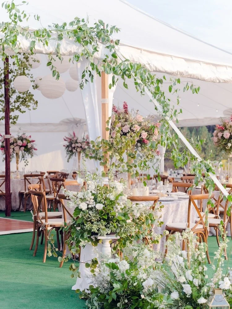Elegant wedding reception setup with round tables and wooden chairs under a white tent, decorated with lush green foliage and white flowers.