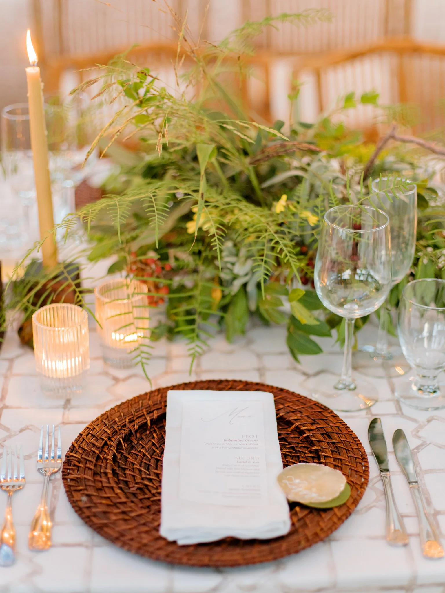 Elegant table setting with a round woven placemat, a white napkin, cutlery, glasses, candles, and a lush green floral centerpiece.