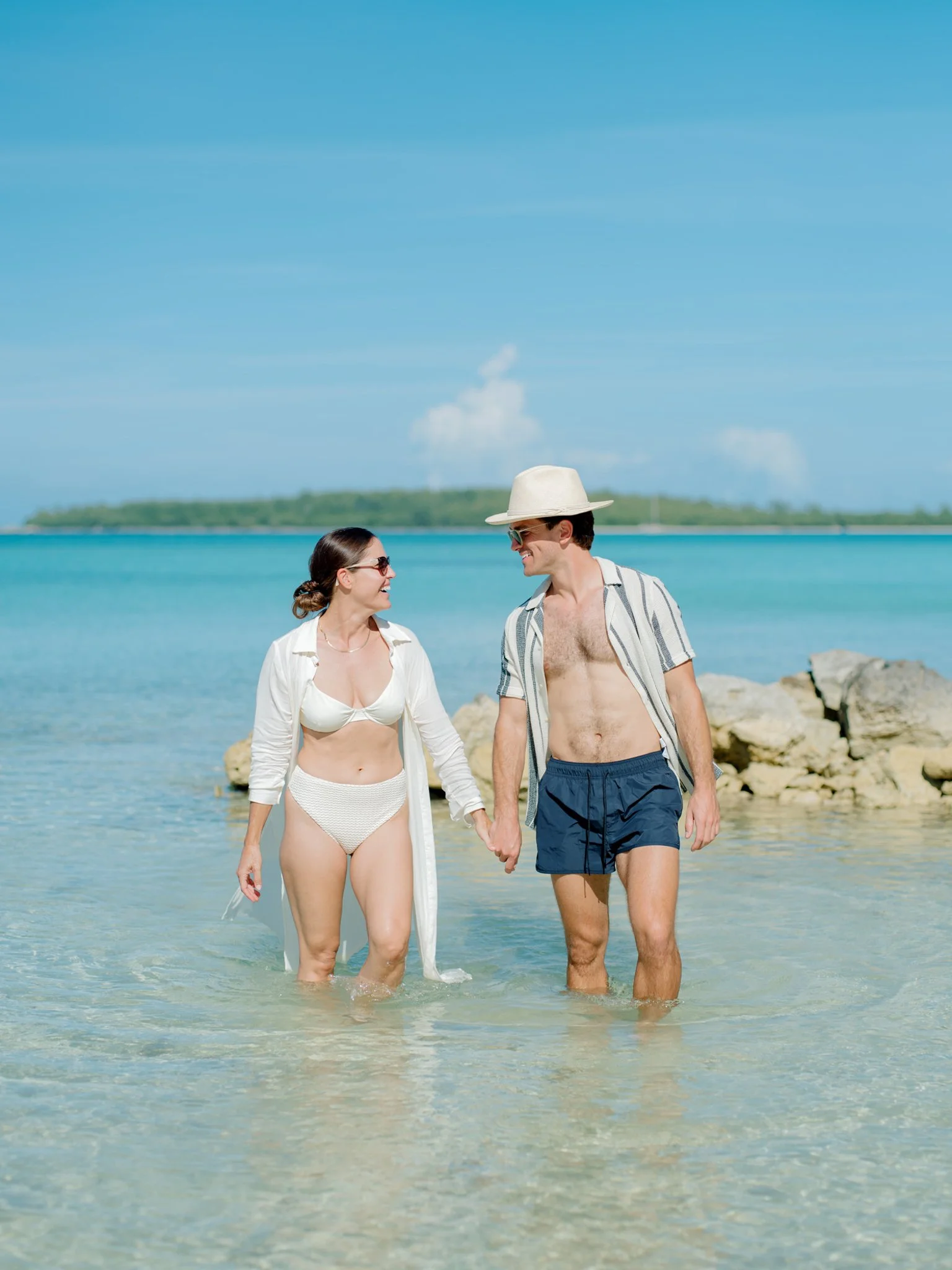 A couple walking hand in hand into the ocean, wearing beach attire, with a clear blue sky and distant land in the background at The Other Side Hotel during their destination wedding in Eleuthera, Bahamas.