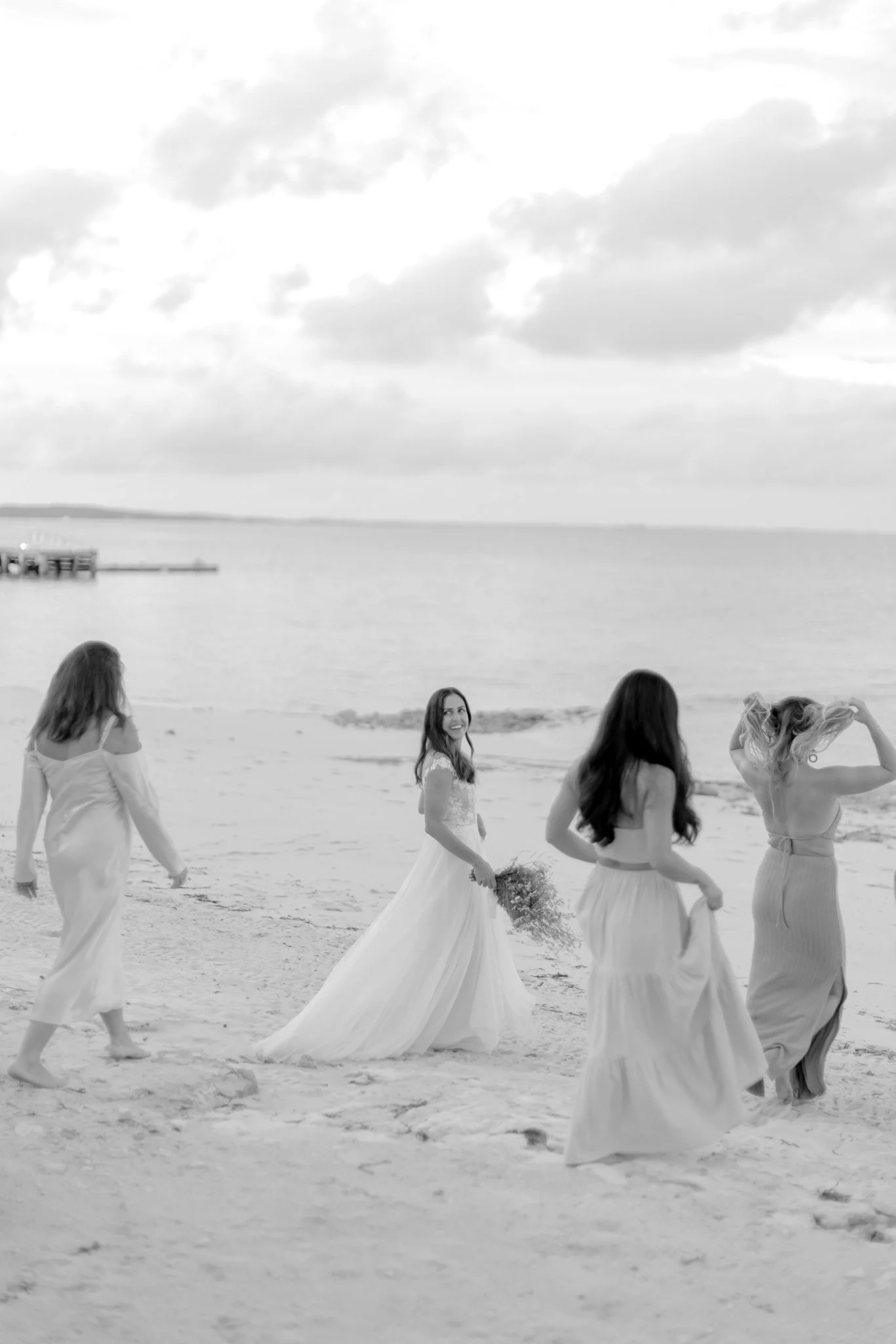 A bride and three bridesmaids walking on a beach, with the bride smiling and holding a bouquet, as the beach, water, and cloudy sky are in the background at The Other Side Hotel Eleuthera during a luxury Bahamas destination wedding.