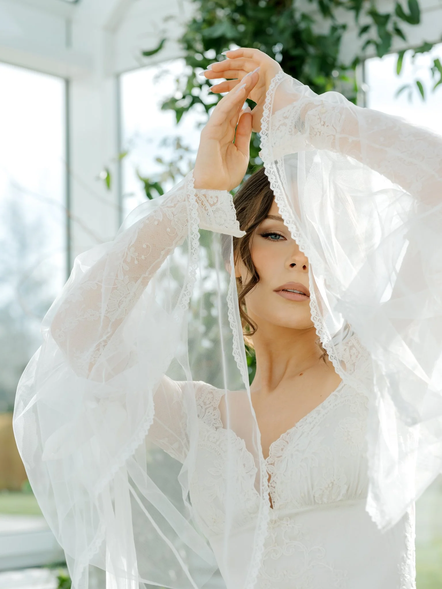 A woman in a sheer white lace dress and veil, her right hand above her head, posing with a greenhouse and greenery behind her at Greenhouse on Harvard near Spokane, Washington
