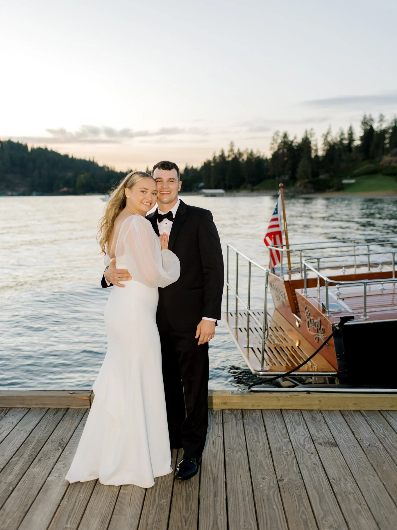 A newlywed couple in wedding attire stands on a wooden dock by a lake, with a boat and trees in the background at sunset at the Hagadone Event Center in Coeur d'Alene, Idaho.