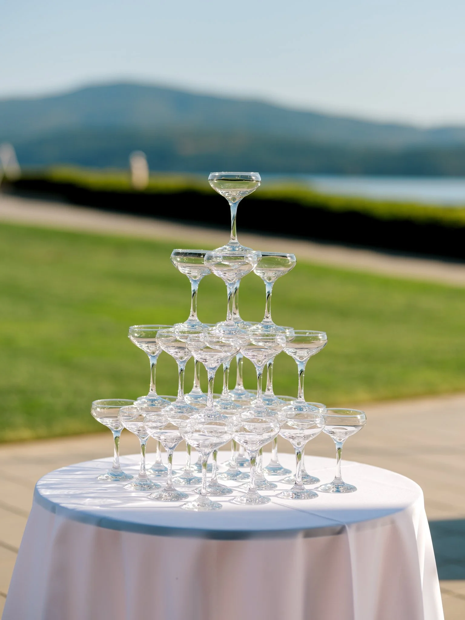 Stacked champagne glasses arranged in a pyramid on a white tablecloth with a blurred natural background and mountains in the distance.