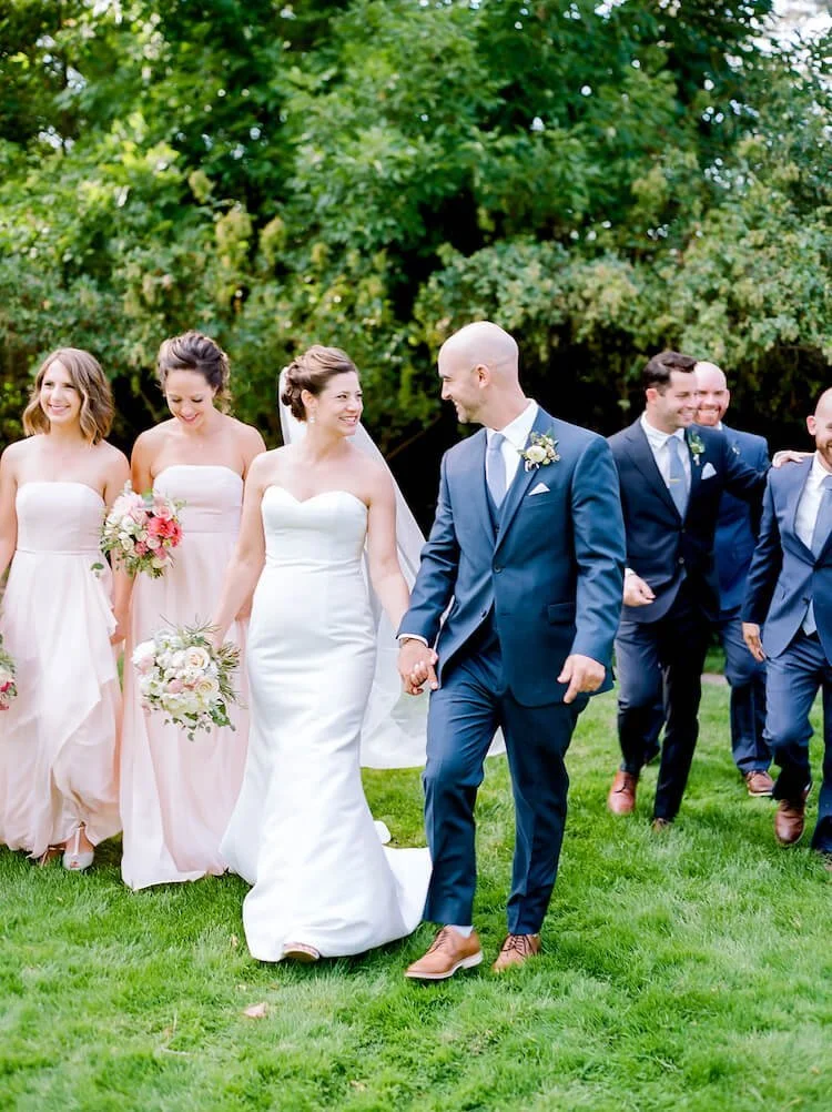 Bride and groom walking outdoors on grass, smiling and holding hands, surrounded by bridesmaids and groomsmen, with trees in the background.
