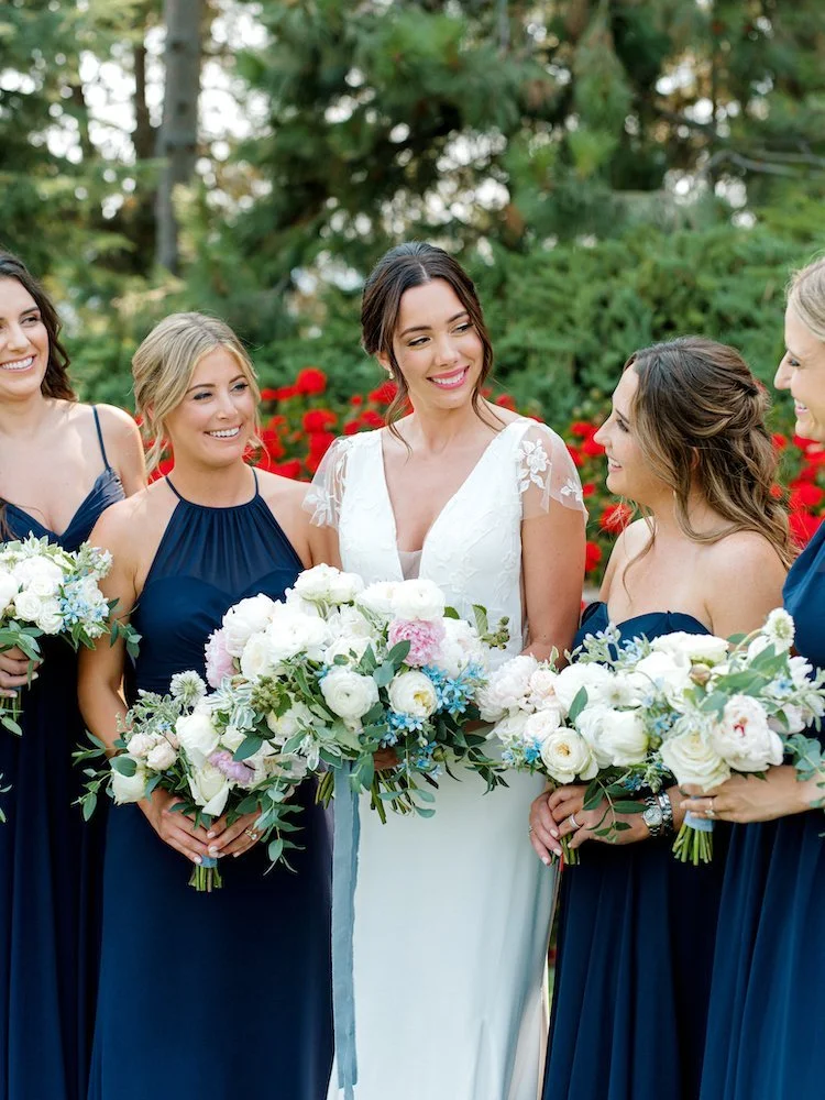 Bride in a white wedding dress with lace cap sleeves, surrounded by four bridesmaids in navy blue dresses, holding bouquets of white and pink flowers, outdoors with greenery and red flowers in the background.