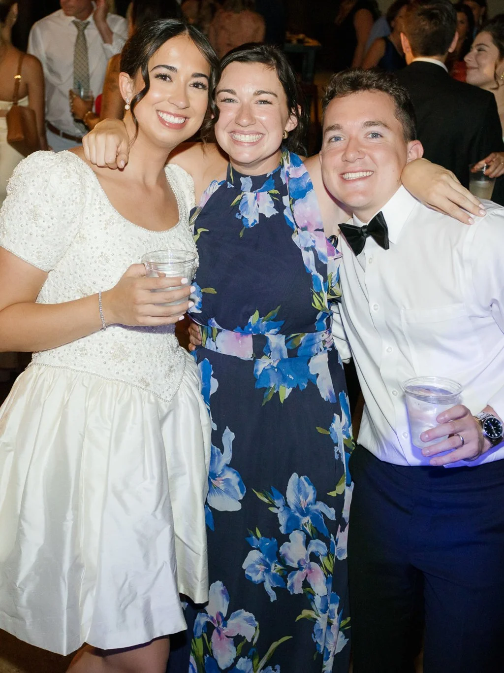 Three people smiling at a party, two women and one man, dressed in formal attire, holding cups, with many other people in the background.