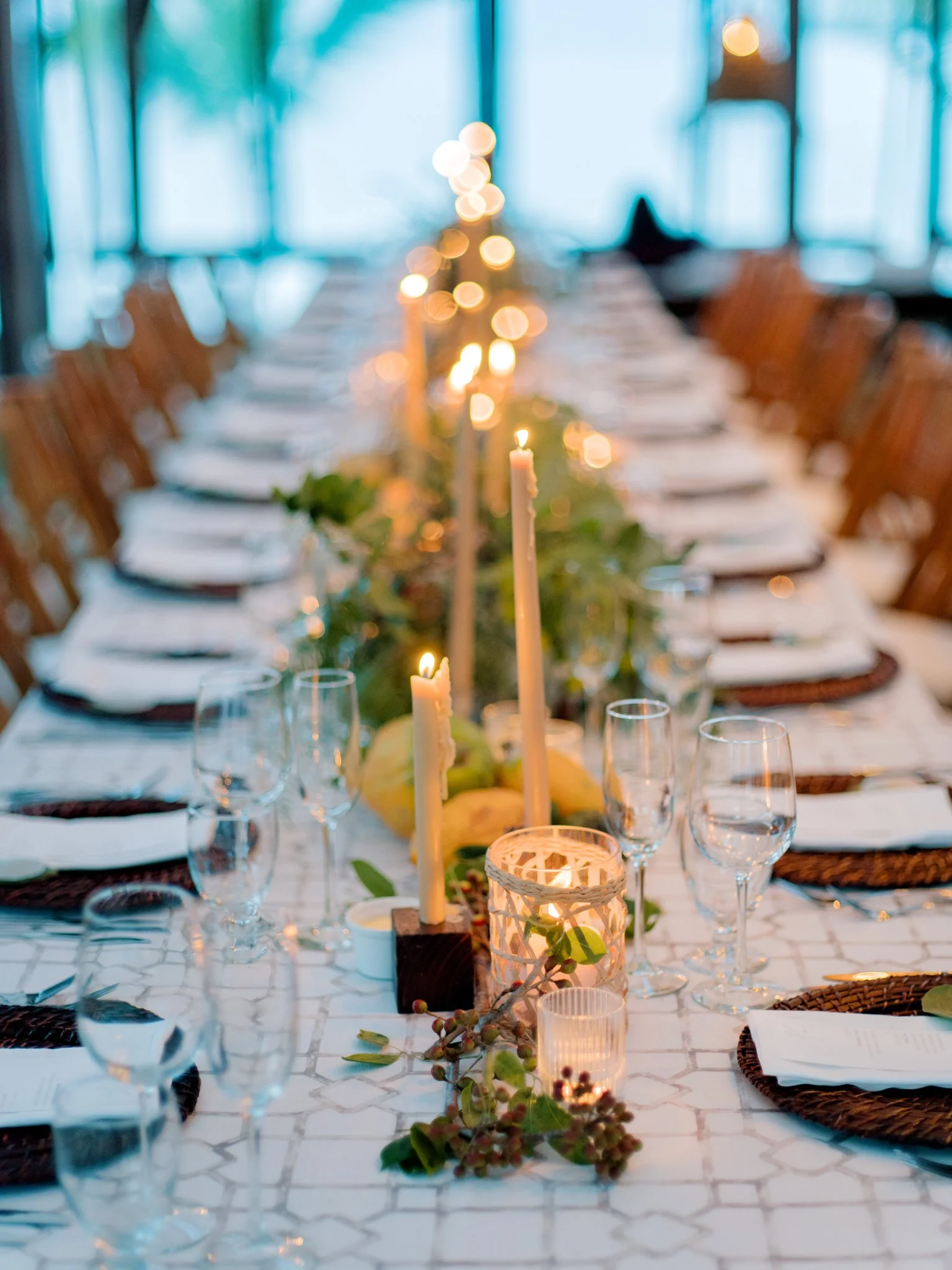 A long dinner table decorated with candles and greenery, set for a celebration or event in a bright room with large windows.
