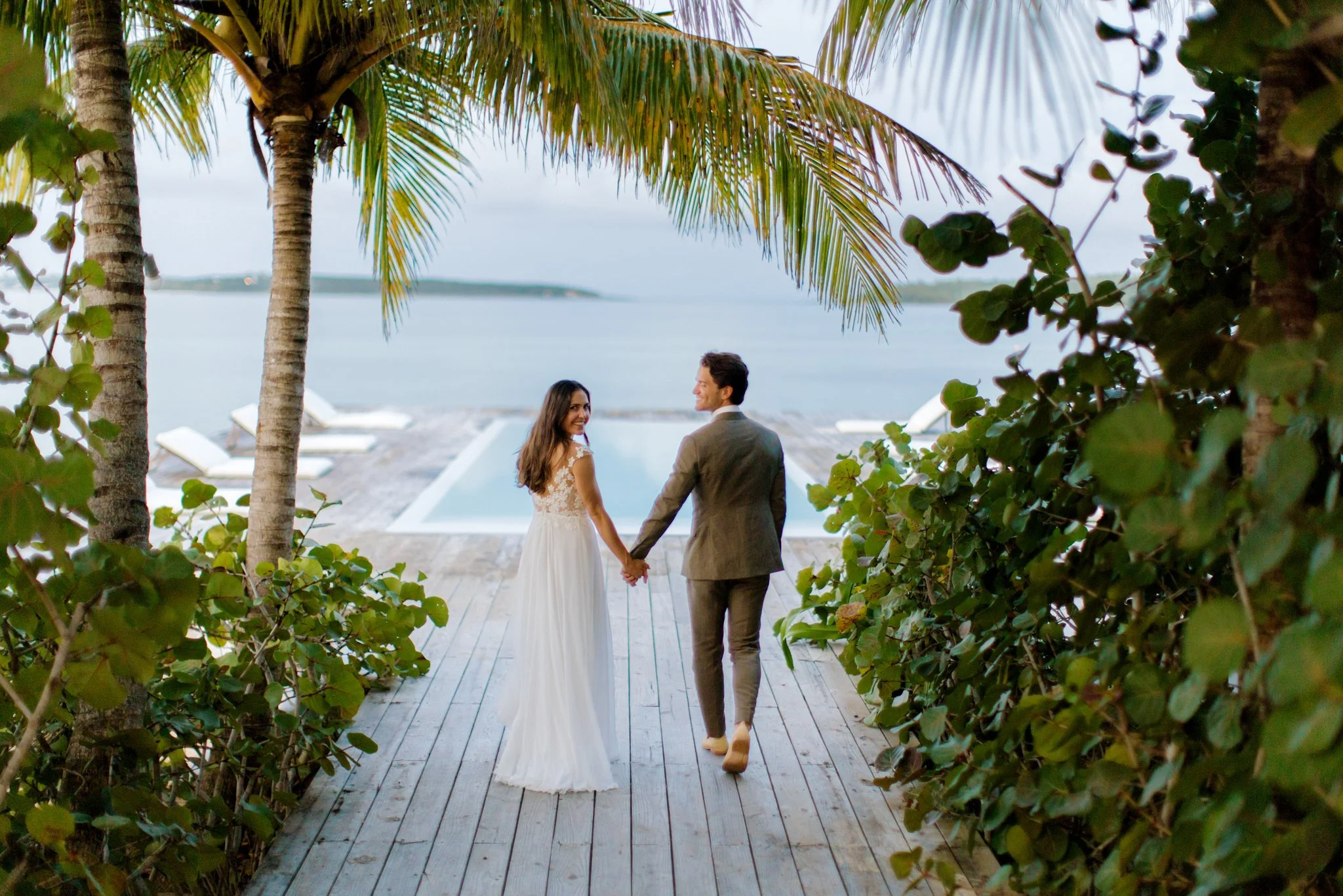 A bride and groom holding hands, walking on a wooden deck towards a pool and ocean view, surrounded by palm trees and green bushes at The Other Side Hotel Eleuthera during a luxury Bahamas destination wedding.