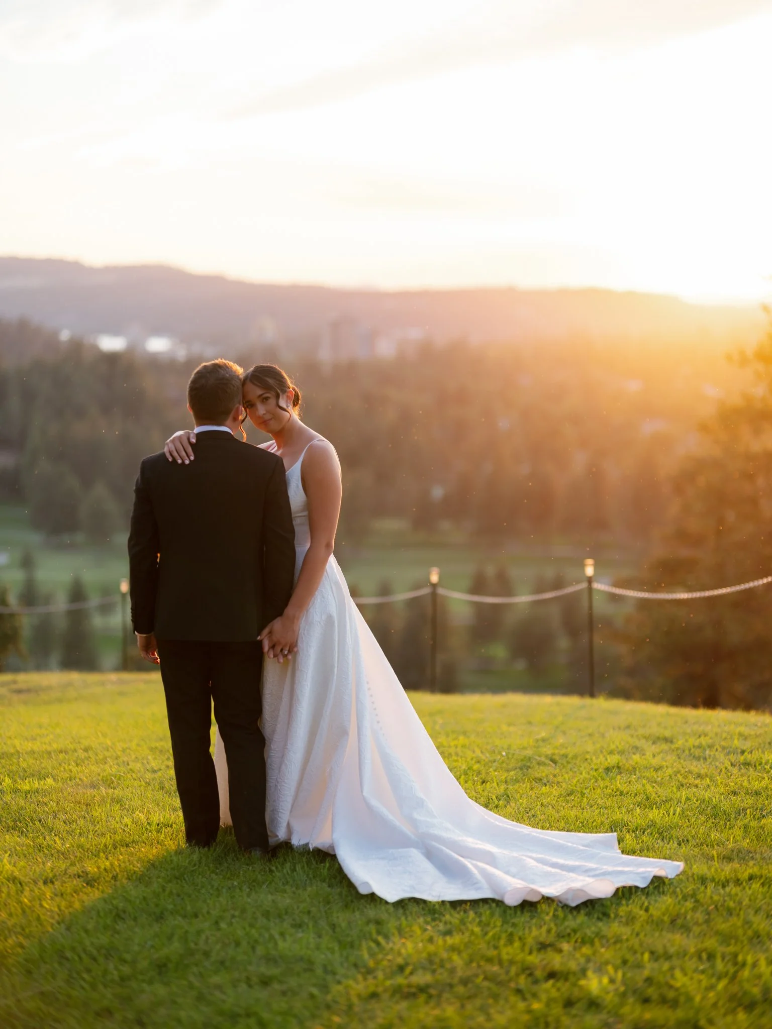 Bride and groom embrace on a grassy field during sunset, with the bride in a white gown and the groom in a black tuxedo at Elk Point Wedding Venue overlooking Lake Coeur d'Alene.