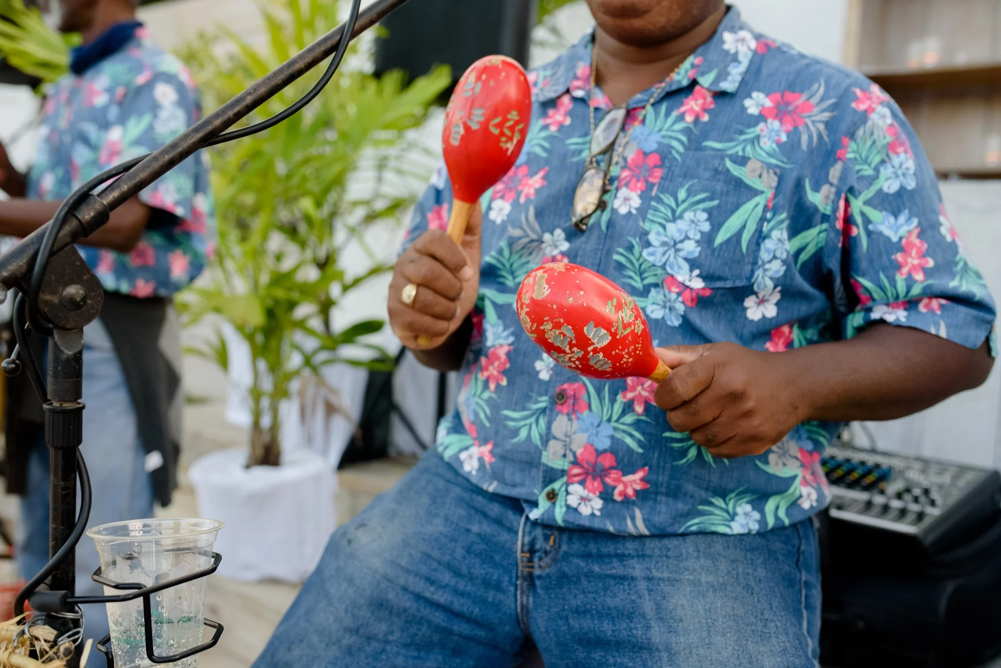 Man holding two red maracas with gold leaf design, wearing a blue floral shirt, at an outdoor event.