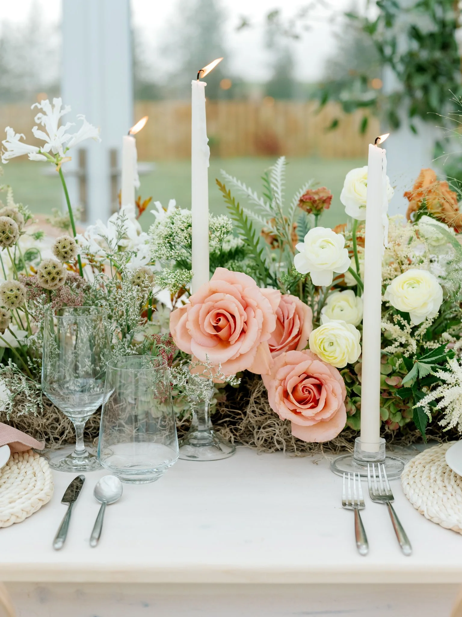 Elegant table setting with a floral centerpiece of pink and white roses, greenery, and candles in a bright room with a window and outdoor view.
