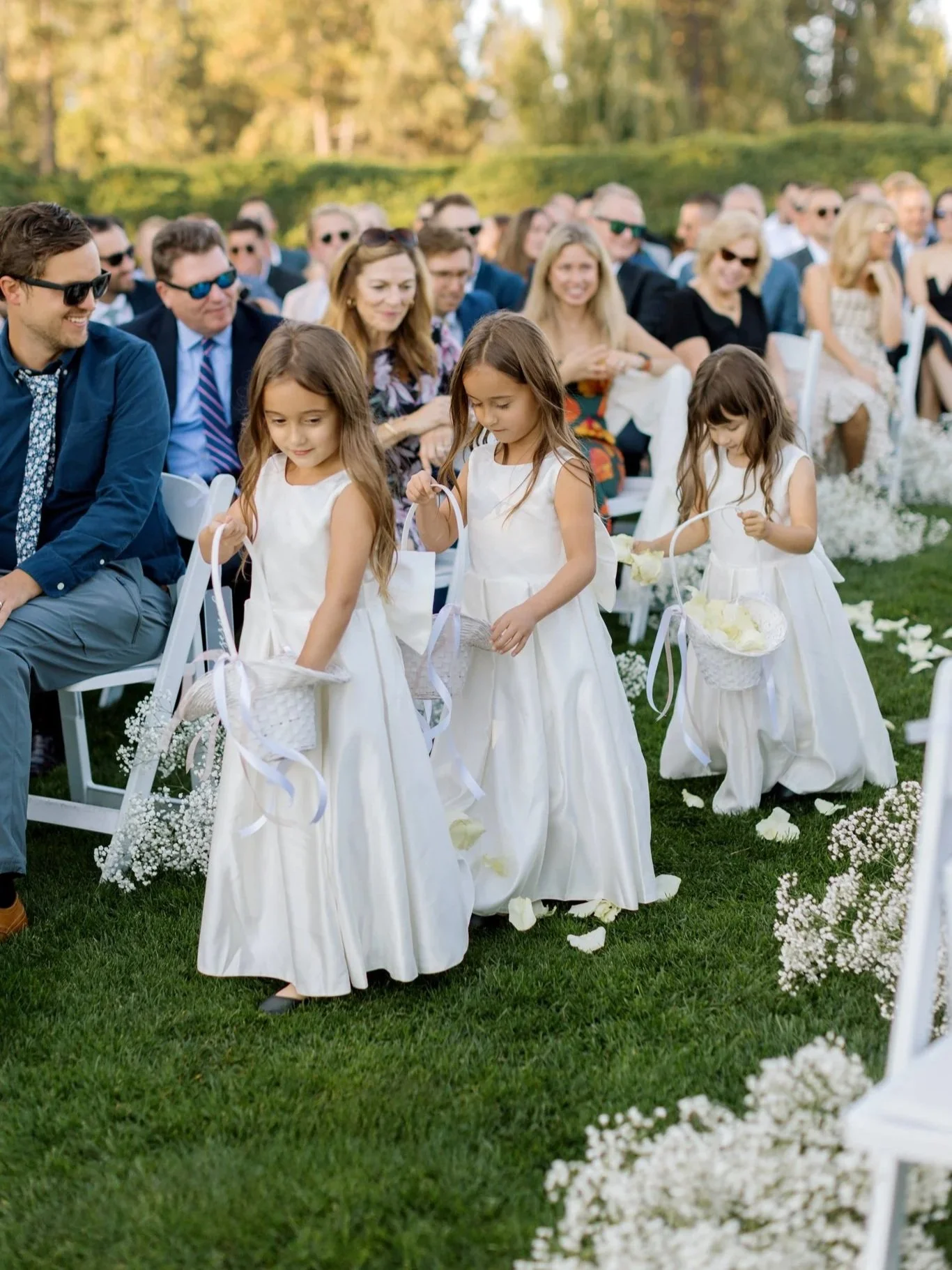 Three young flower girls in white dresses scattering petals as they walk down the aisle during a lakeside ceremony in Coeur d’Alene.