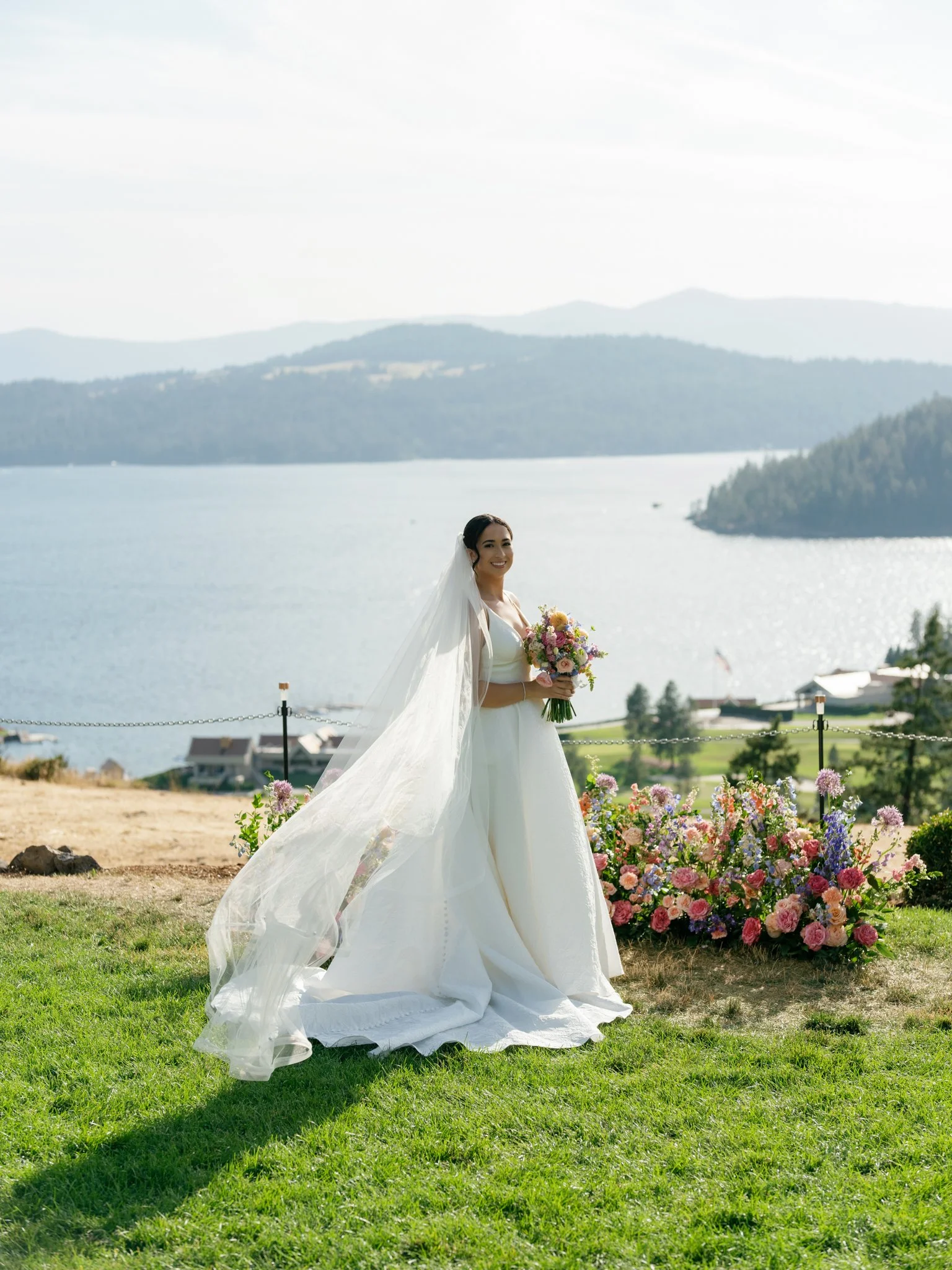 Bride in white wedding dress holding a bouquet outdoors near a lake, with flowers and landscape in the background at Elk Point wedding venue above Lake Coeur d’Alene.