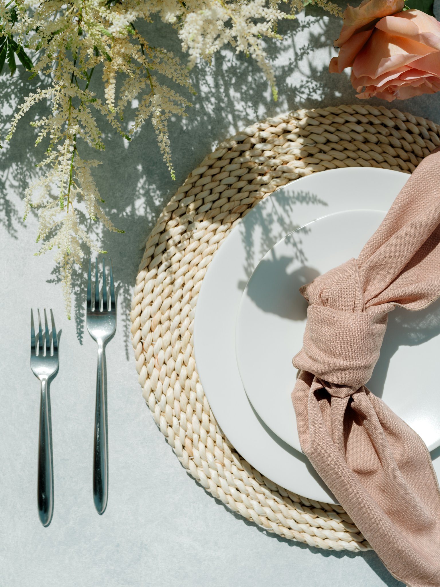 A table setting with a white plate, beige napkin, and two silver forks on a woven placemat, with a floral arrangement on a white surface at Greenhouse on Harvard near Spokane, Washington