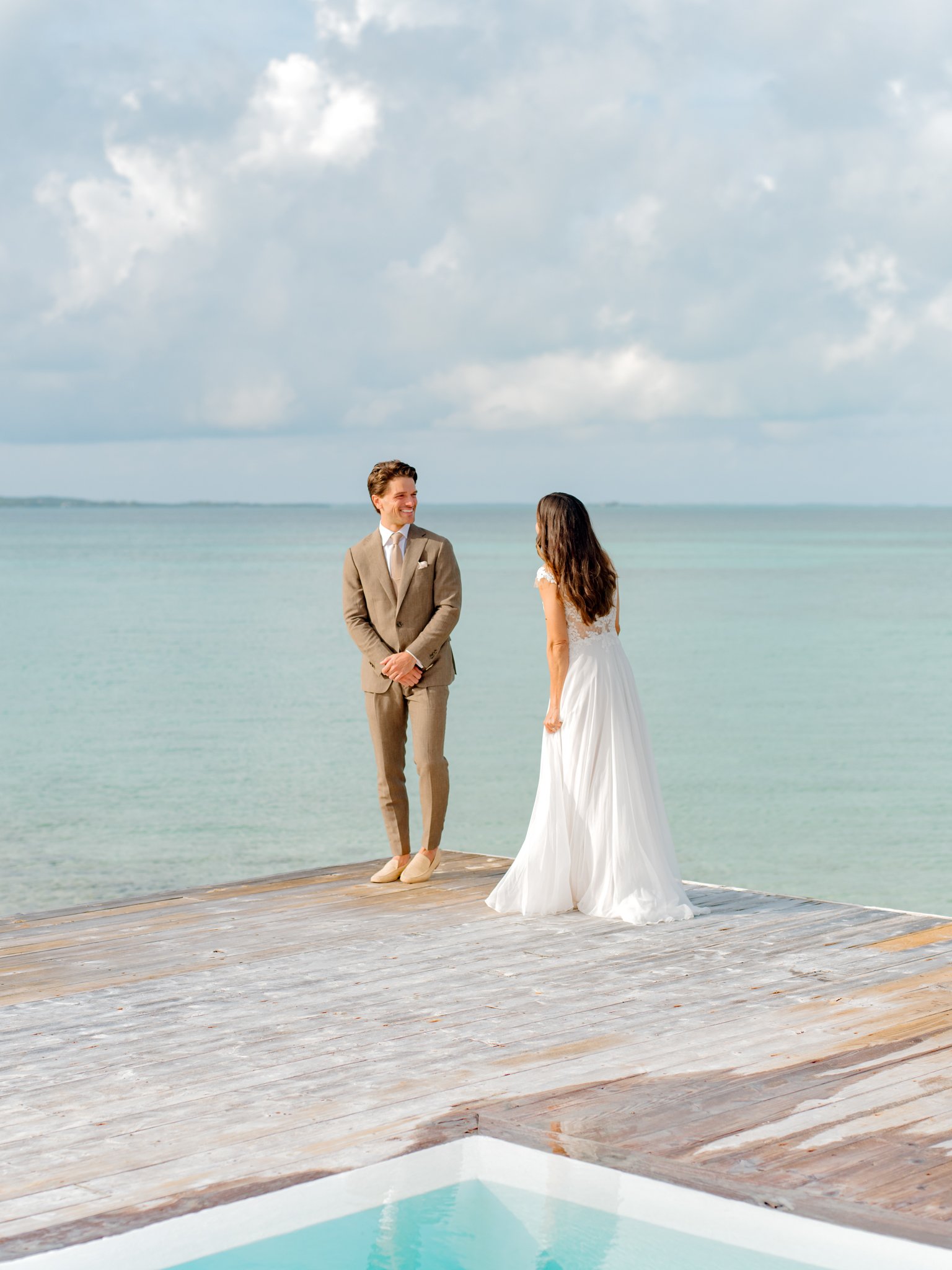 A bride and groom standing on a wooden dock by the water, facing each other, with the sky and clouds in the background at The Other Side Hotel Eleuthera during a luxury Bahamas destination wedding.
