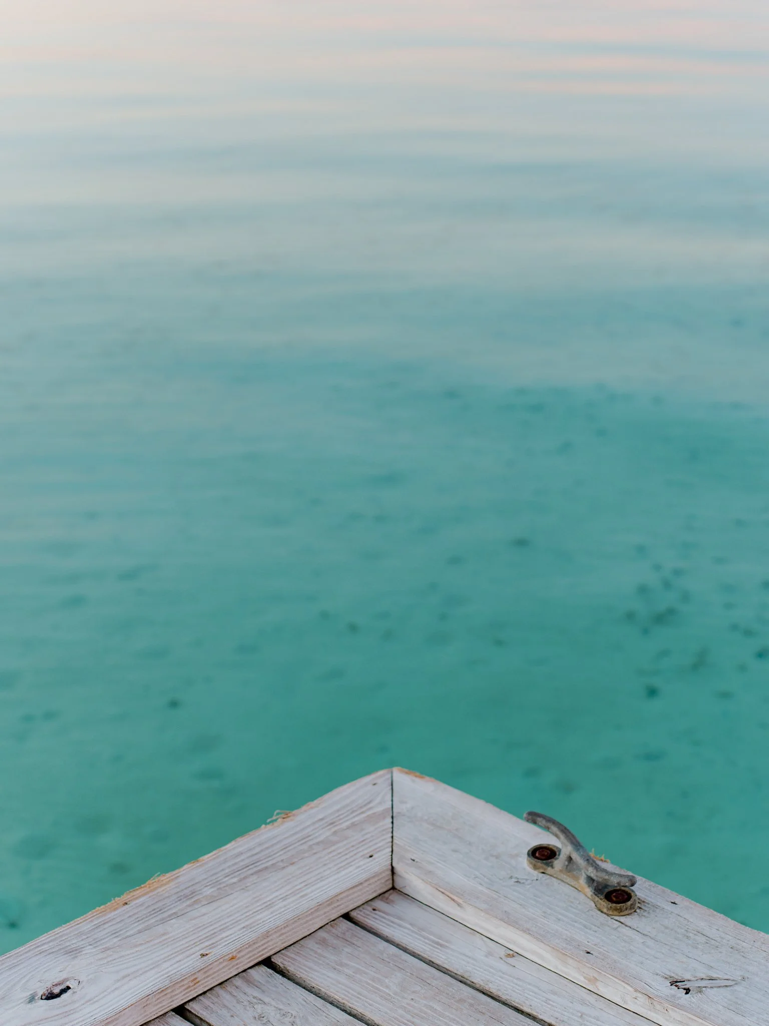 Close-up of a wooden dock corner with rusted metal hardware, overlooking clear turquoise water and a calm ocean horizon.
