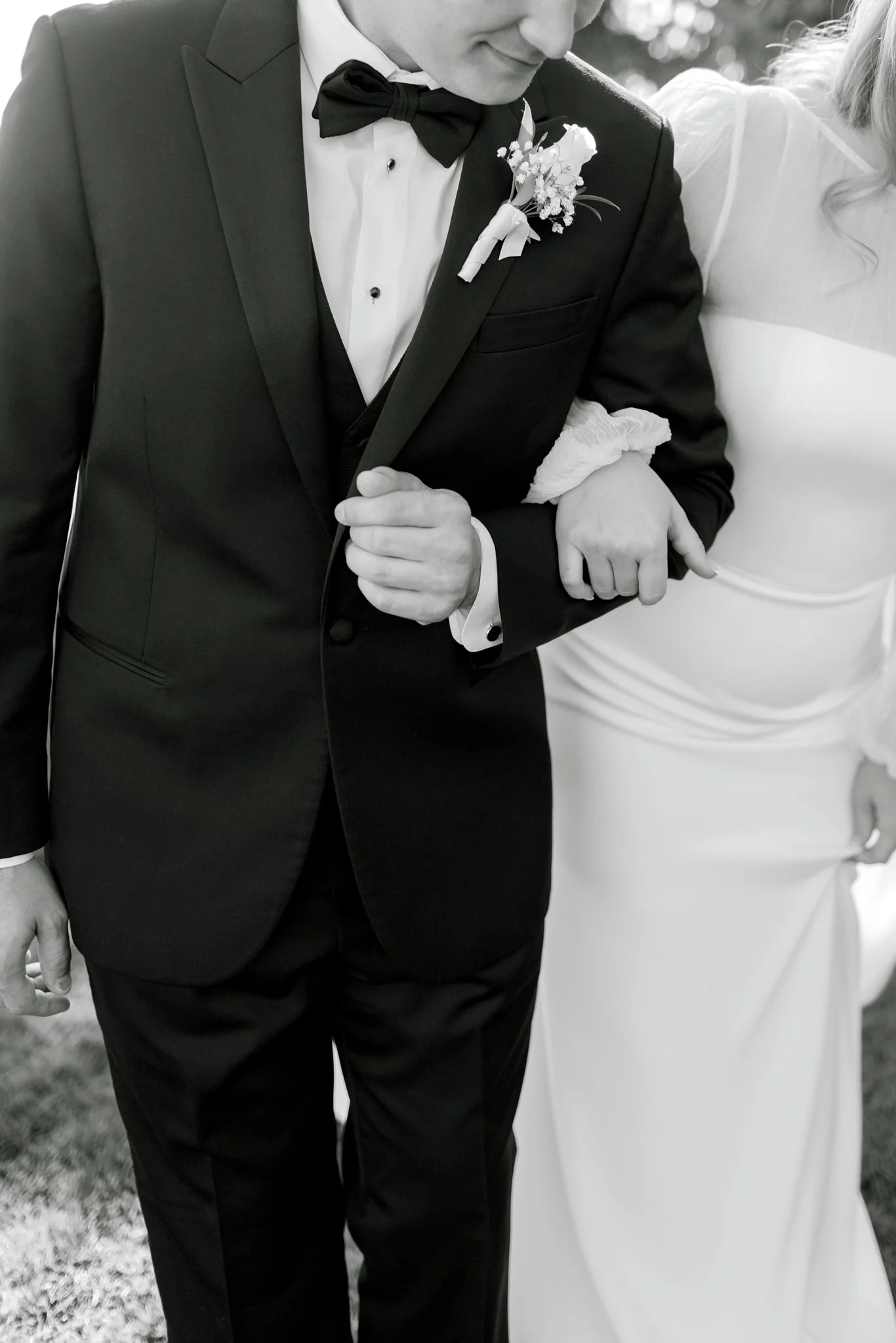 A groom in a tuxedo holding the hand of a bride, both standing close together at a wedding.