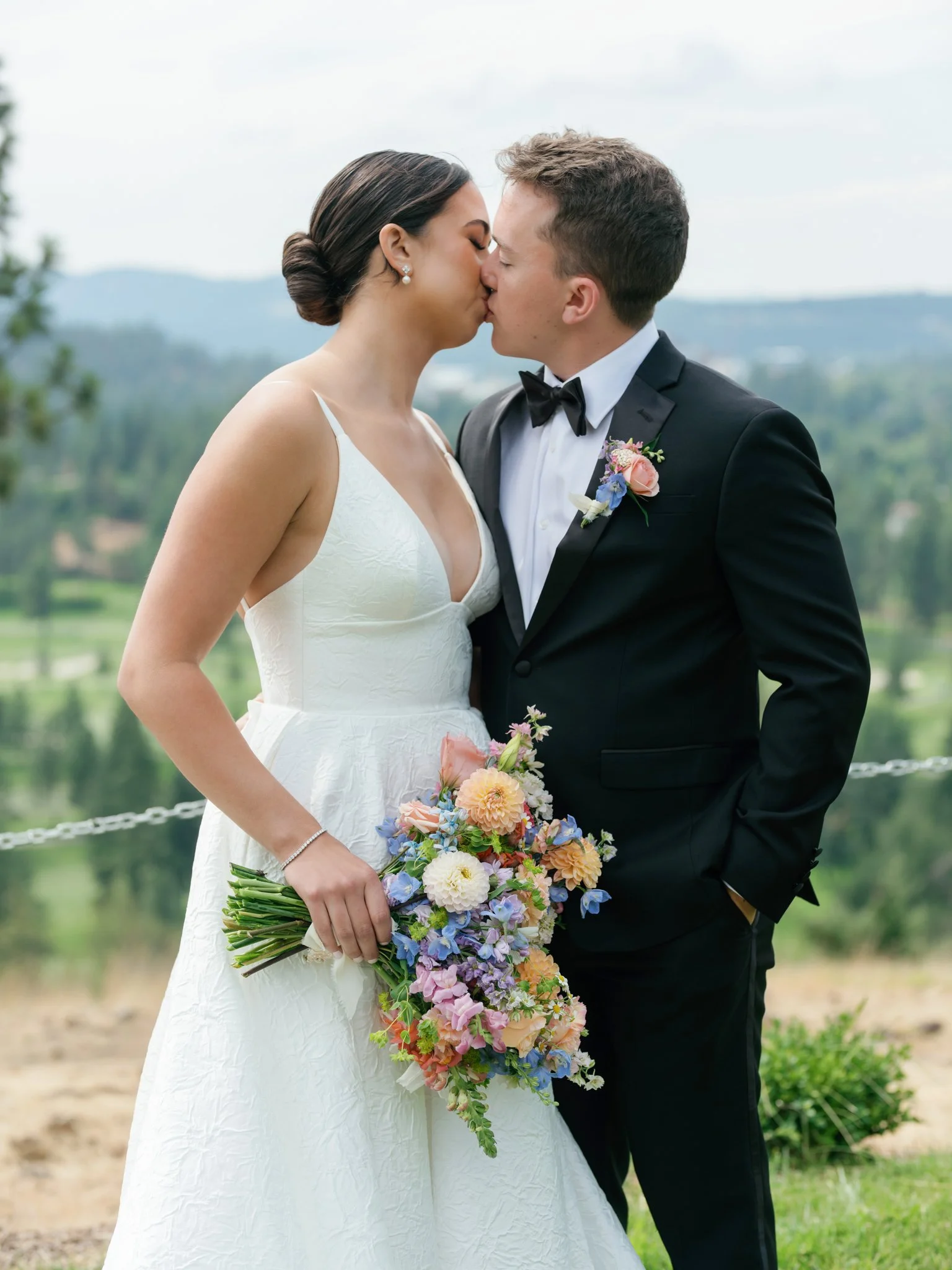 A bride and groom kissing outdoors, with the bride holding a colorful bouquet of flowers, and the groom dressed in a black tuxedo with a bow tie and boutonniere at Elk Point wedding above Lake Coeur d’Alene.