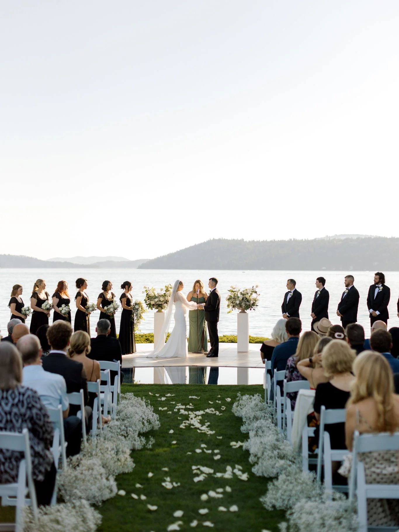 Wide ceremony view of the couple exchanging vows with their wedding party and Lake Coeur d’Alene in the background.