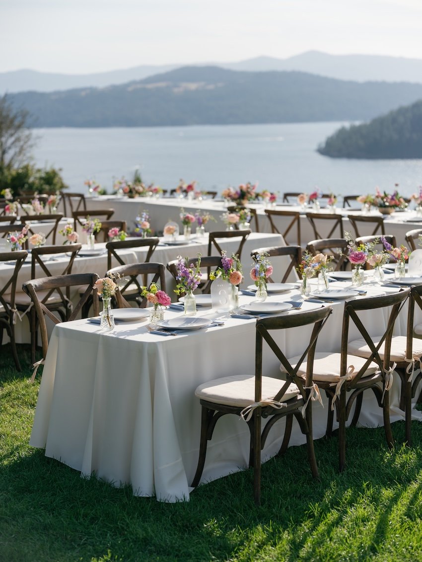 Outdoor banquet tables decorated with white tablecloths, floral centerpieces, and table settings, overlooking a lake and mountains in the distance.