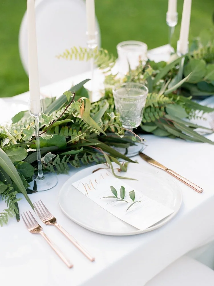 Elegant outdoor table setting with a white tablecloth, greenery centerpiece, rose gold cutlery, clear glassware, candles, and a place card with greenery, suggesting a wedding or special event.