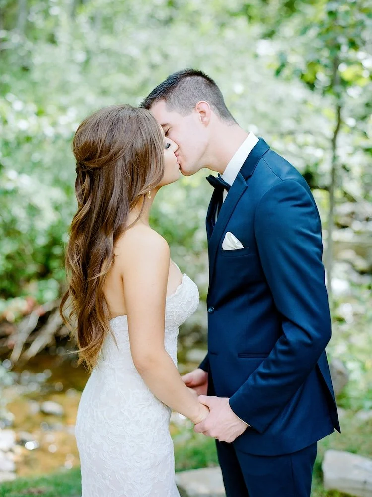 A bride and groom sharing a kiss outdoors, holding hands, with a blurred green forest background.