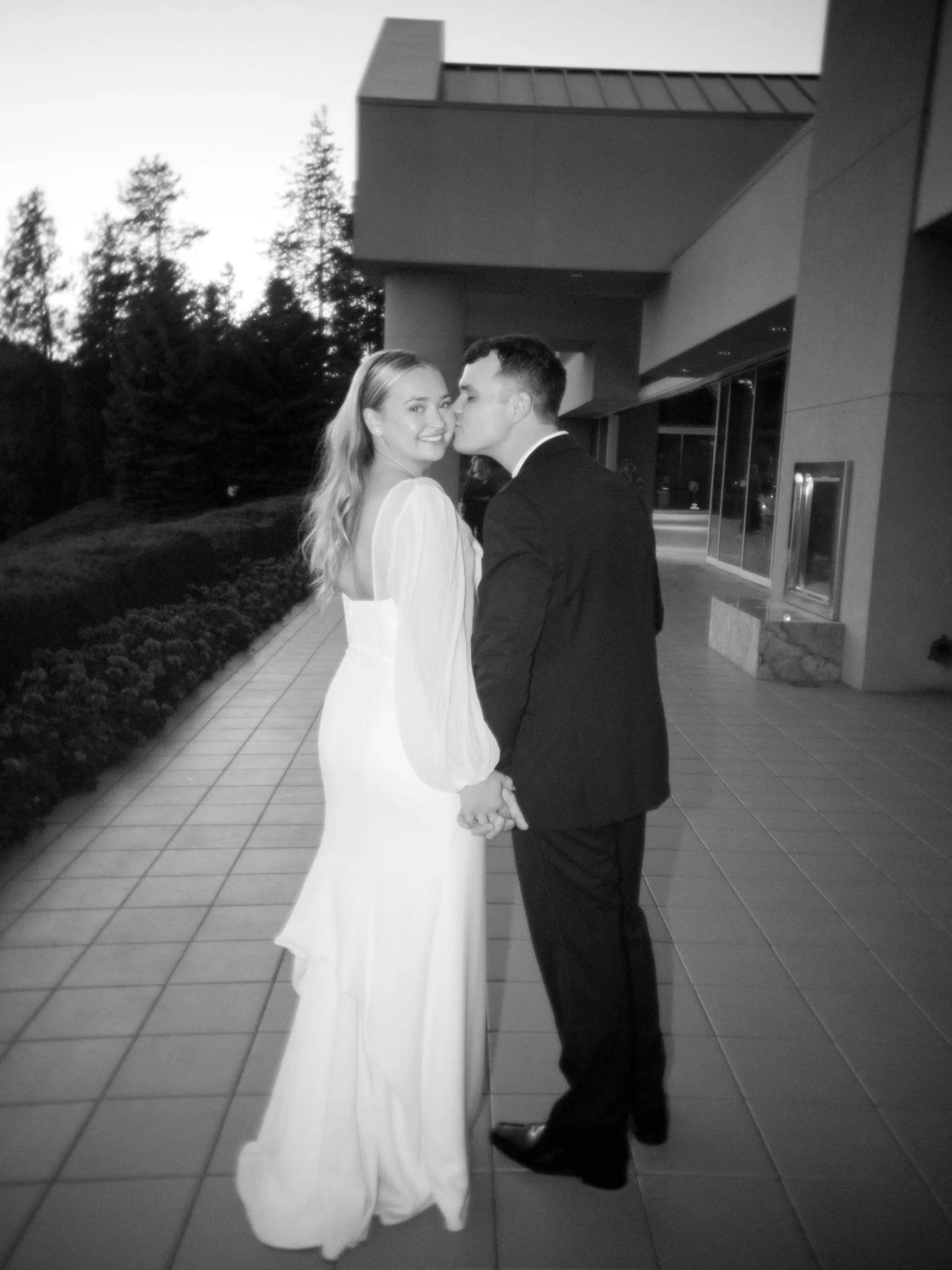 A bride and groom holding hands, standing outside a modern building at sunset, with trees in the background. The bride is smiling at the camera, wearing a long white dress with sheer sleeves, and the groom is kissing her on the cheek, dressed in a dark suit.