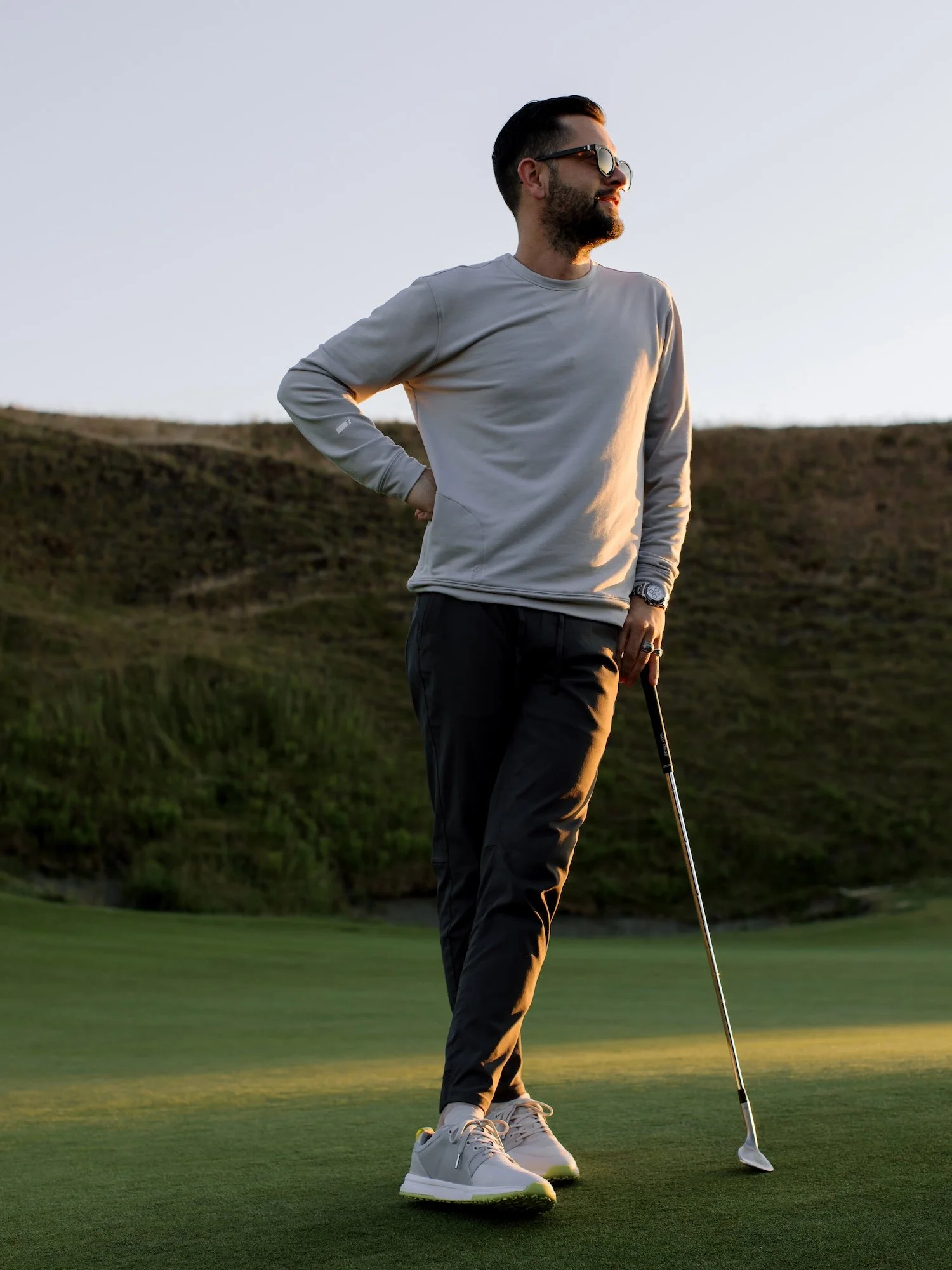 A man standing on a golf course during sunset, holding a golf club, wearing sunglasses, a light gray long-sleeve shirt, dark pants, and light gray sneakers.