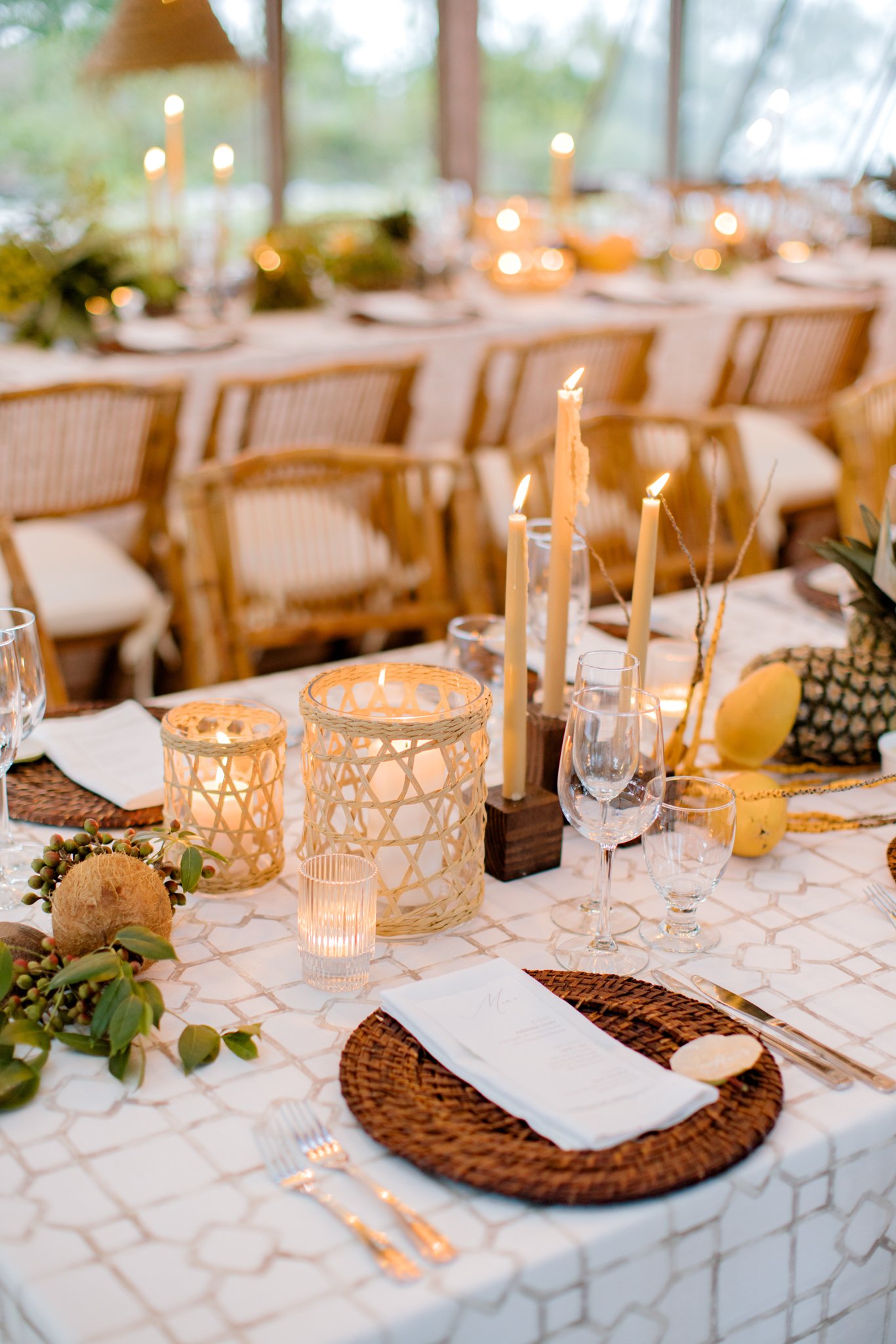 Decorated dining table with candles, wicker lamps, pineapple, and greenery, set for a meal in a bright, airy room with large windows.