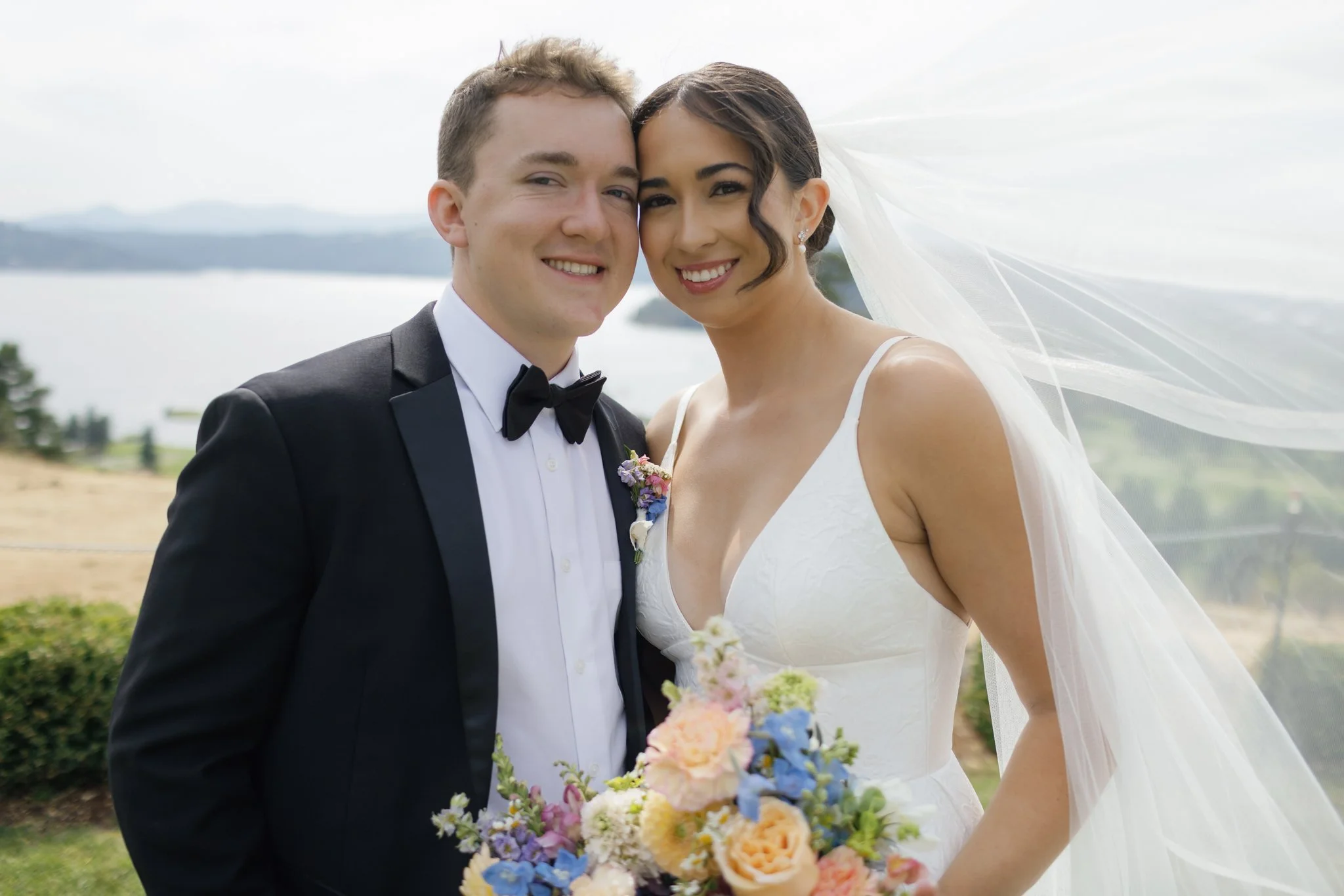 A newlywed couple smiling outdoors with a lake and trees in the background, the bride holding a bouquet of colorful flowers at Elk Point mountaintop wedding.