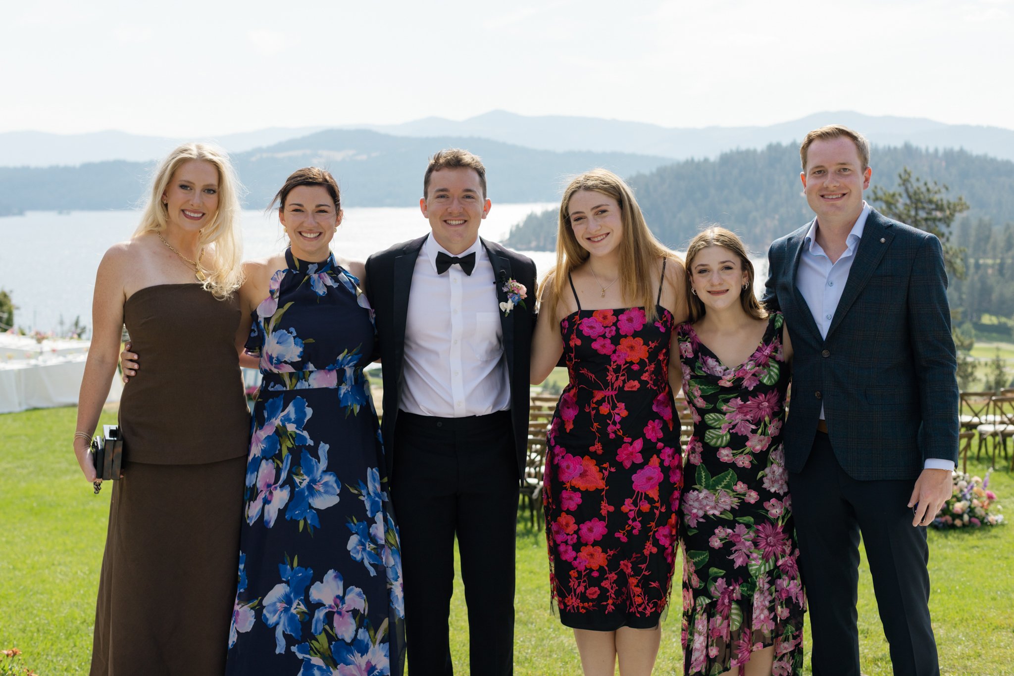 Group of seven people dressed in formal attire, posing outdoors on a grassy area with a lake and mountains in the background. The group includes women in dresses and men in suits, smiling for the camera.