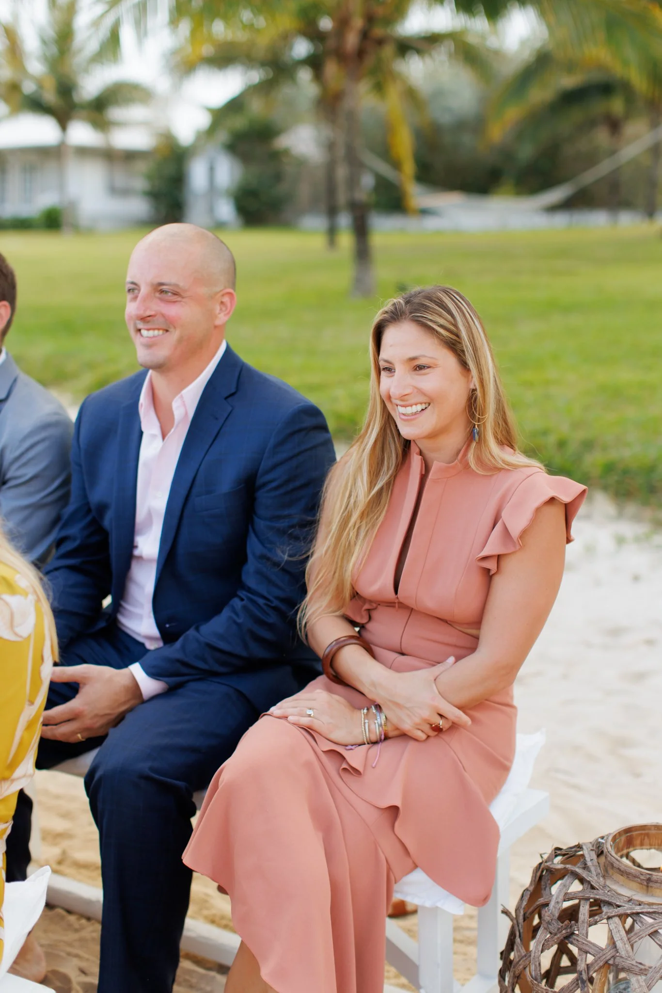 A smiling woman with long blonde hair in a peach dress sitting on a white chair outdoors, with a man in a navy suit next to her, at a beachside event with palm trees and a grassy area in the background.