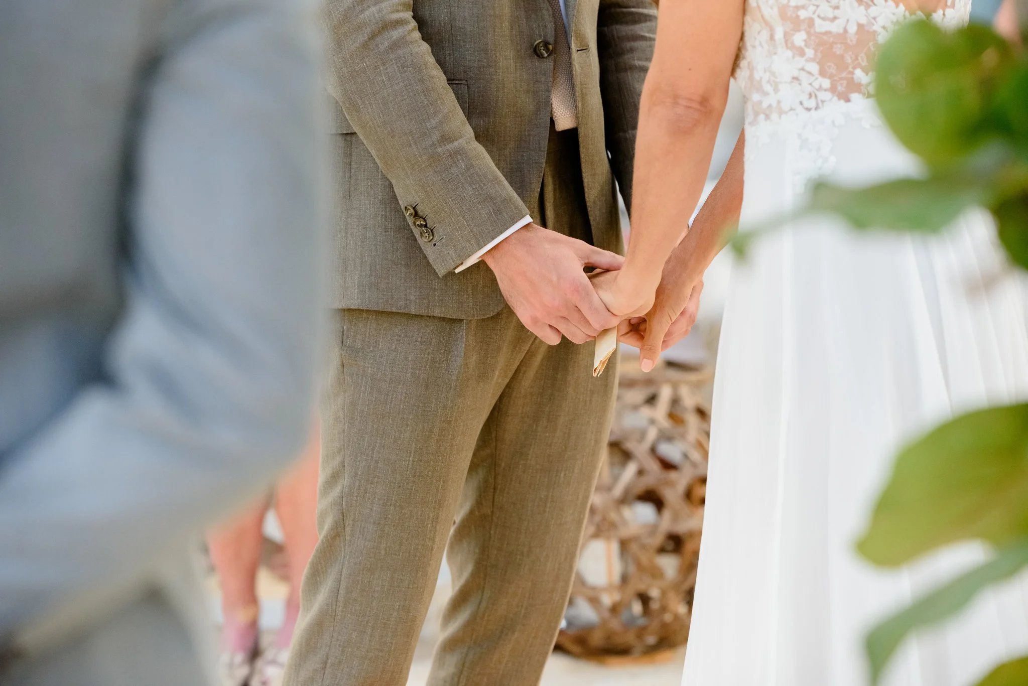 Close-up of a couple holding hands during a wedding ceremony, with the bride wearing a white dress and the groom in a gray suit.