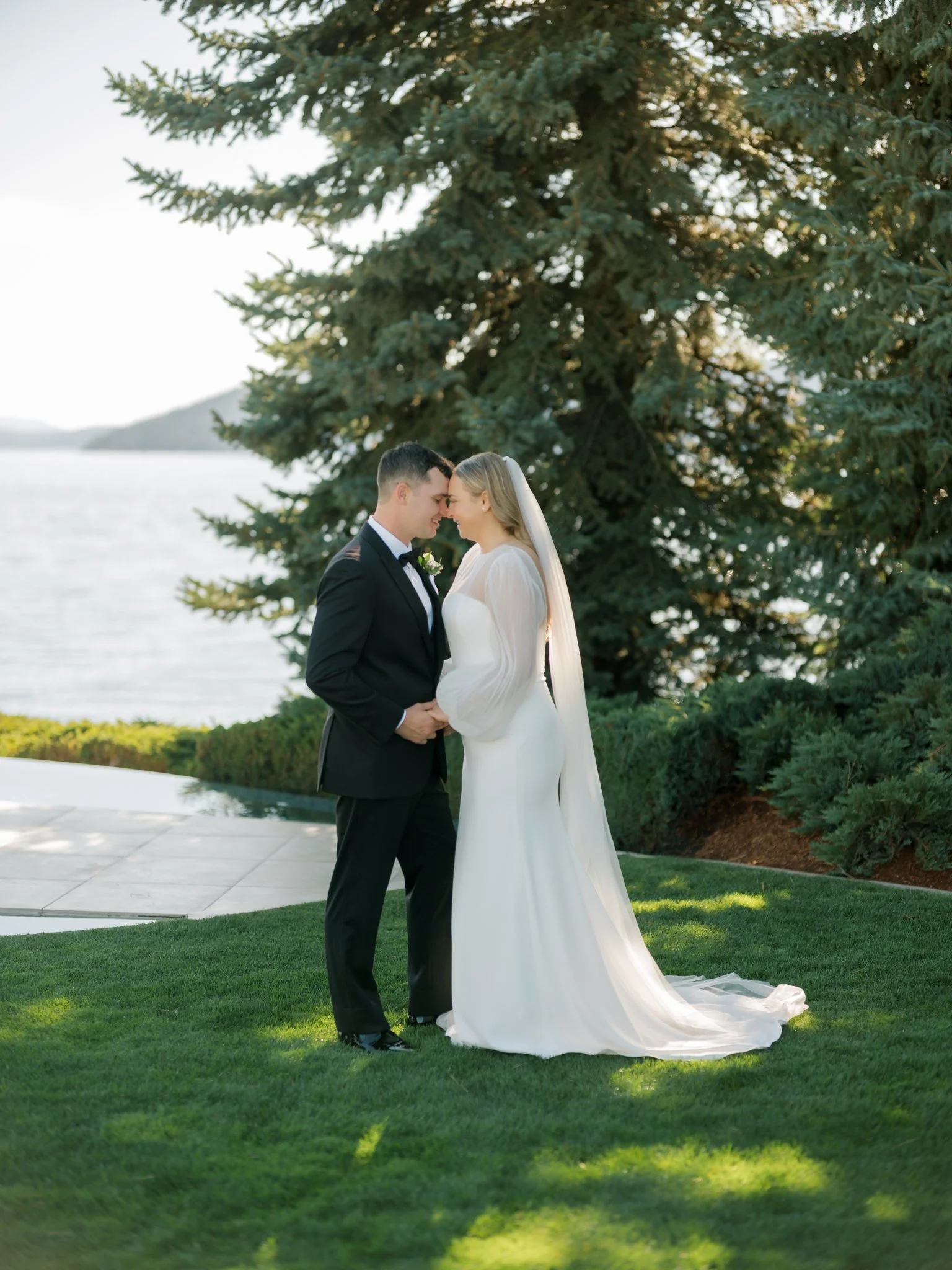 Bride and groom standing close with foreheads touching beneath the pines at their Coeur d’Alene wedding.
