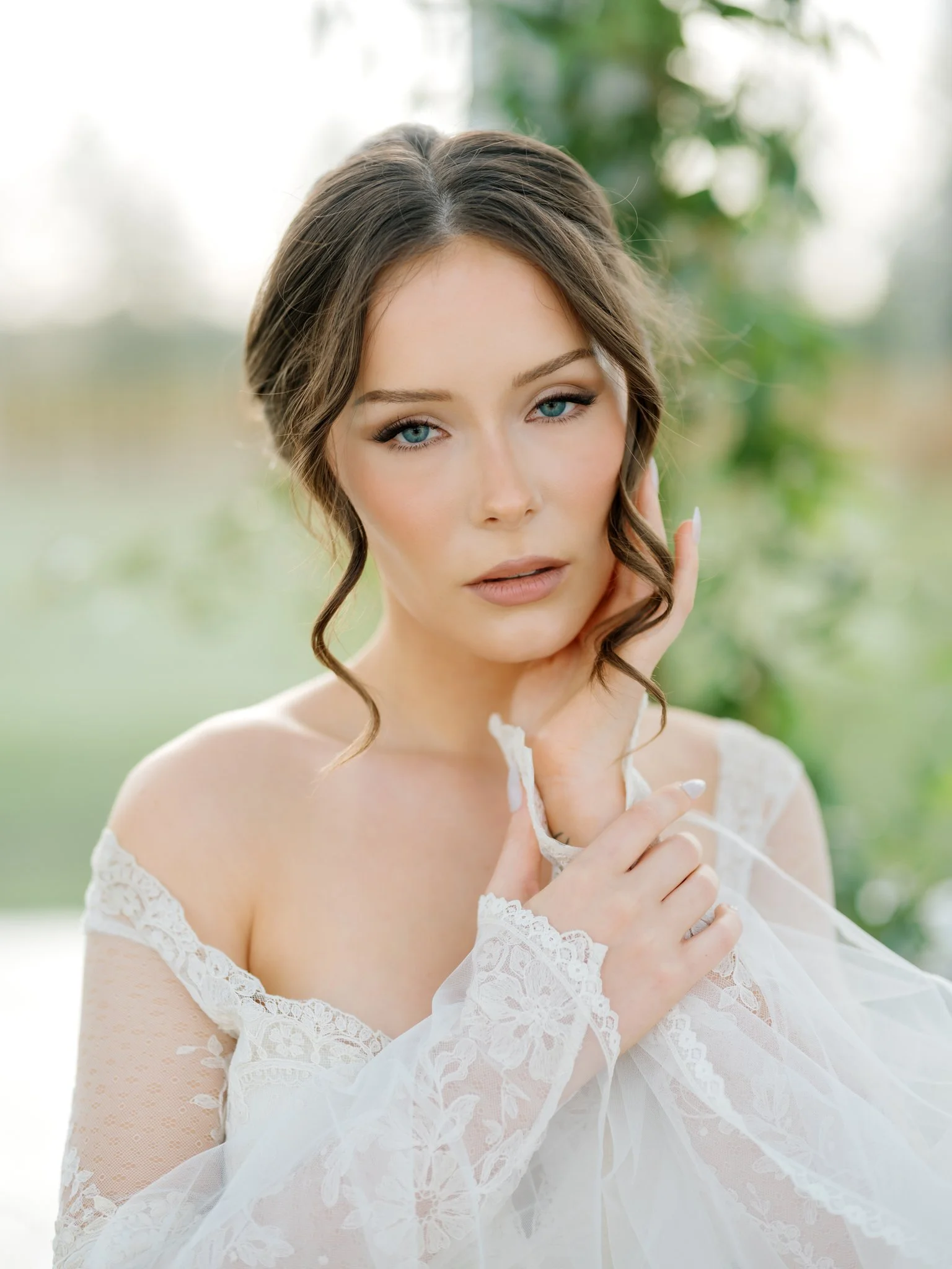 A woman with blue eyes and brown hair styled in loose curls, wearing a white lace dress, outdoors with a blurred green background at Greenhouse on Harvard near Spokane, Washington.