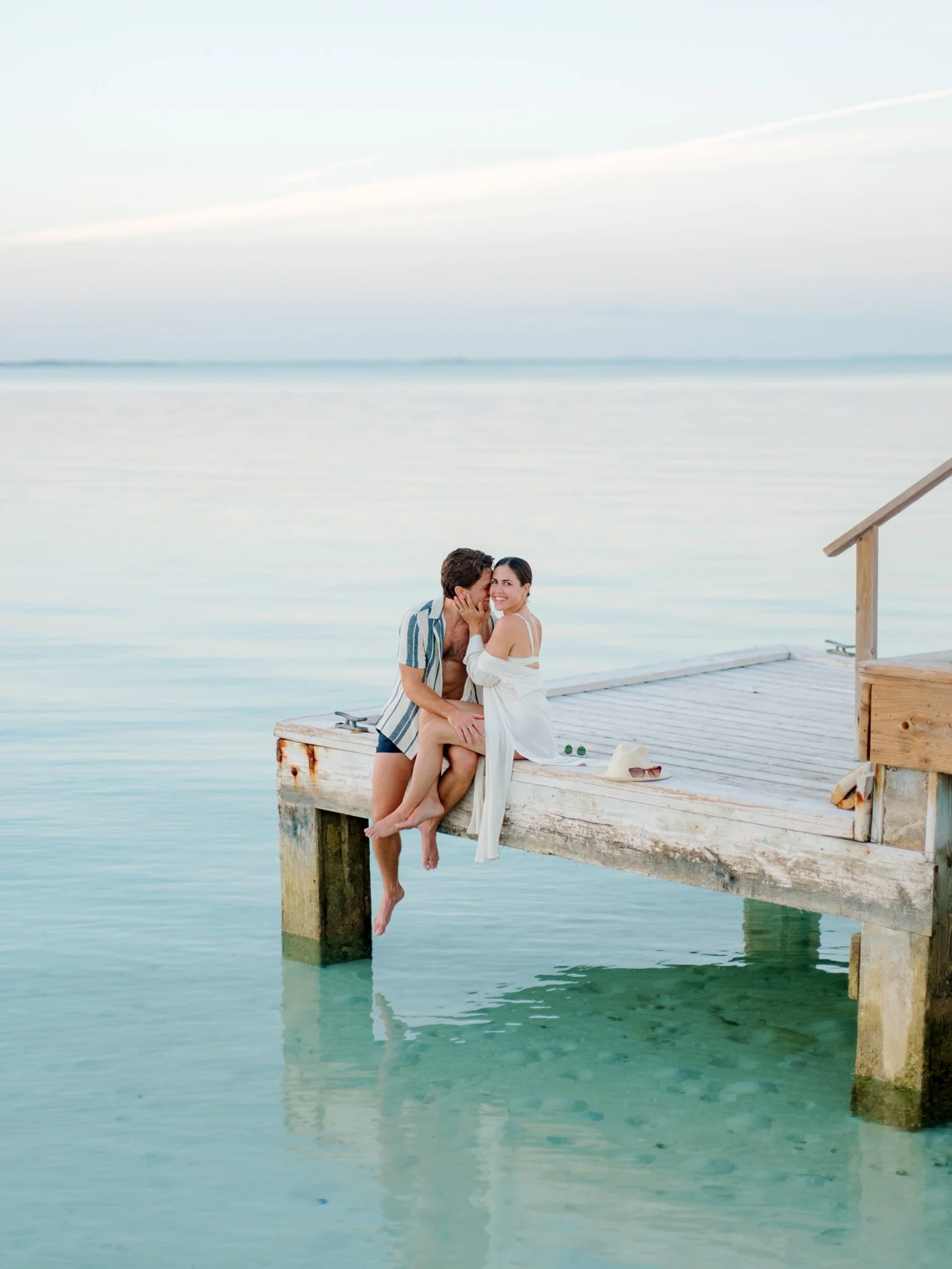 A couple sitting on the edge of a wooden dock over clear water, with the man whispering into the woman's ear while she smiles, wearing casual summer clothes at The Other Side Hotel during their destination wedding in Eleuthera, Bahamas.
