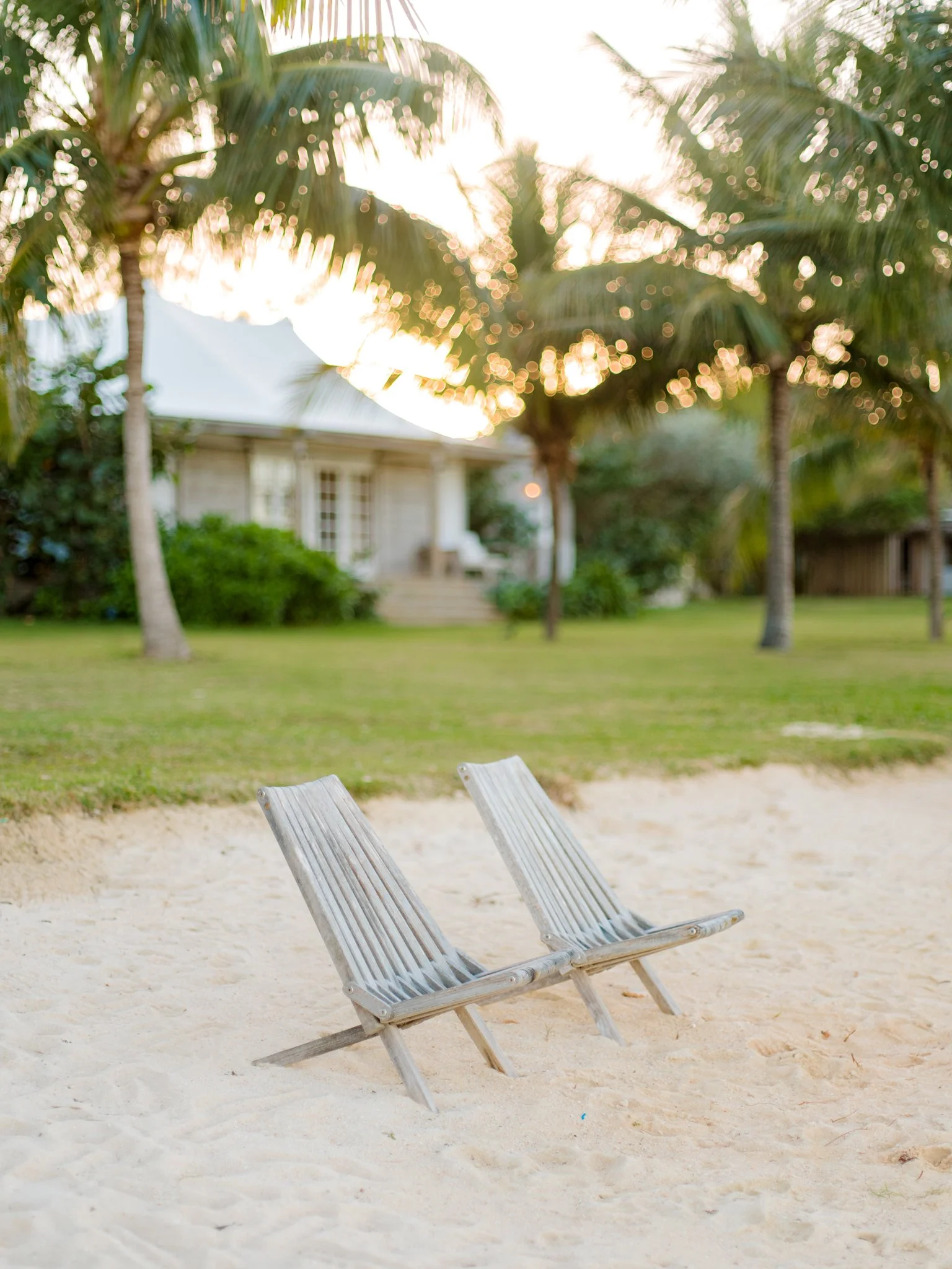 Two wooden lounge chairs on a sandy beach with a grassy yard and house with palm trees in the background, under a setting sun at The Other Side Hotel during their destination wedding in Eleuthera, Bahamas.
