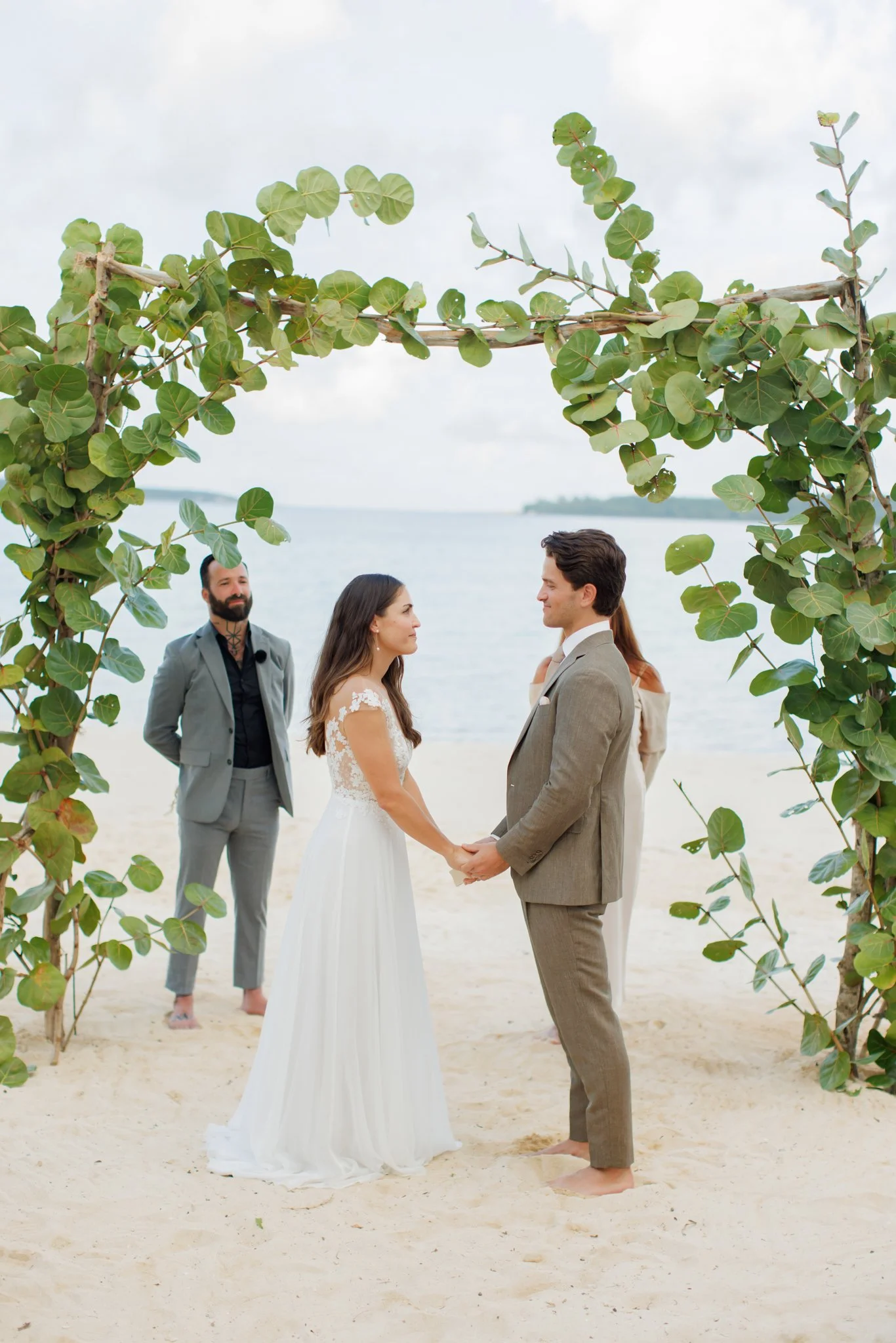 A beach wedding ceremony with a couple holding hands under a leafy arch, with the ocean in the background and an officiant standing behind them at The Other Side Hotel Eleuthera during a luxury Bahamas destination wedding.