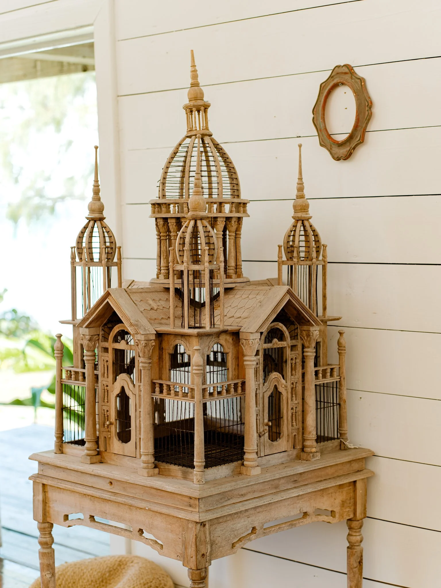 A detailed wooden birdcage with multiple spires and ornate balconies, placed on a small wooden table against a white shiplap wall, with a mirror on the wall.