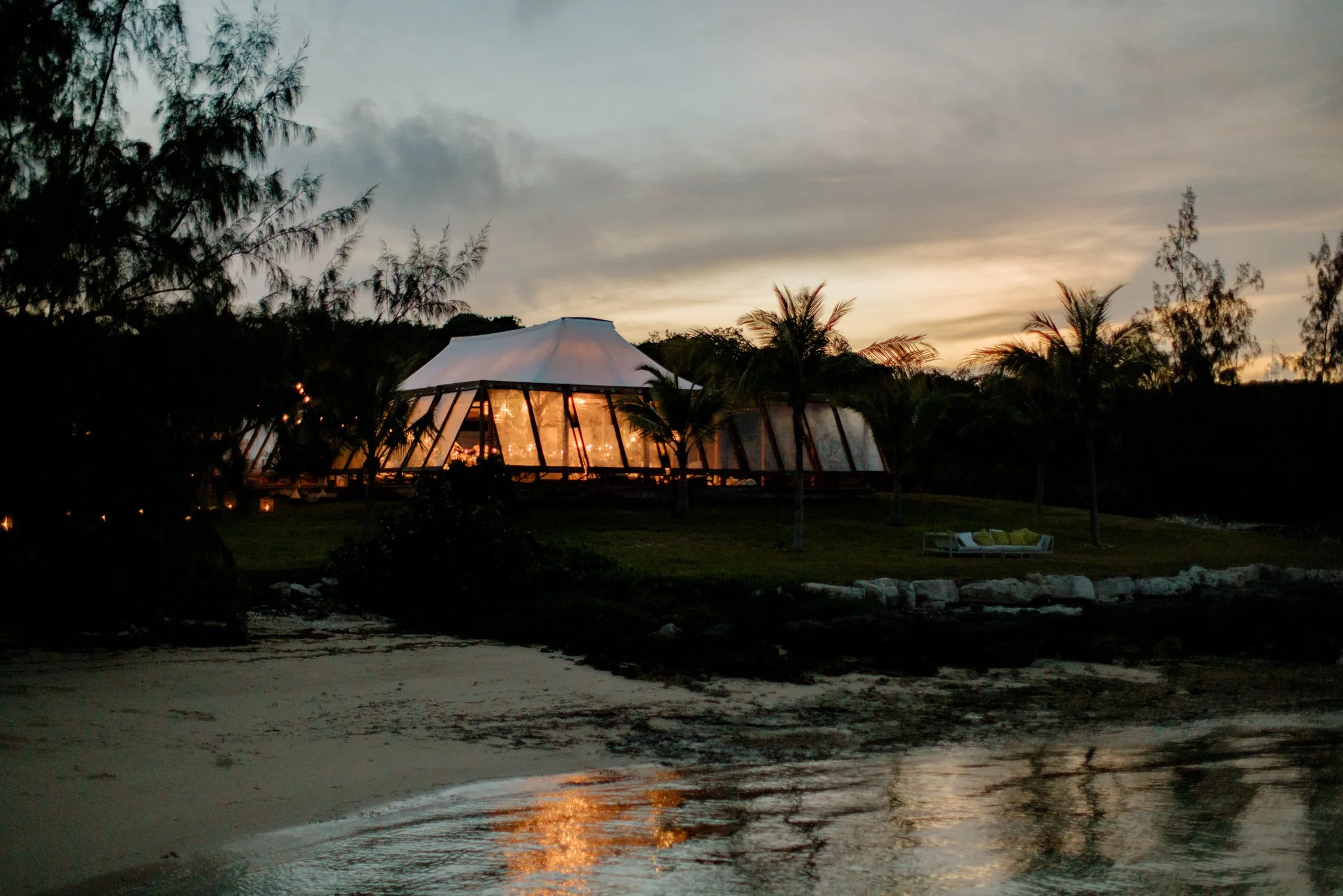 A modern glass building with warm interior lighting is situated by a lake, surrounded by palm trees and greenery during sunset.