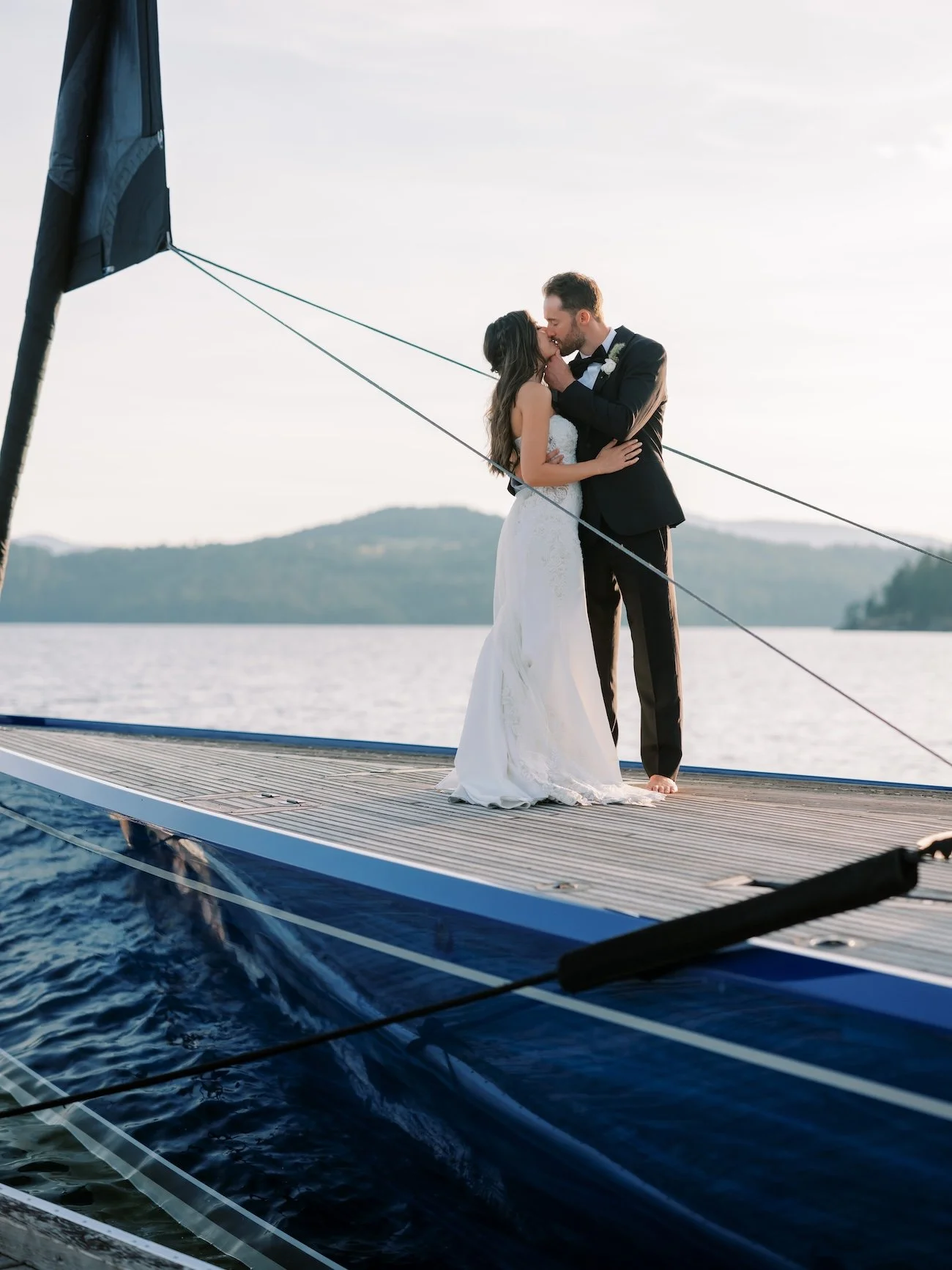 A bride and groom in wedding attire sharing a kiss on a boat floating on the water with a mountain range in the background during sunset.
