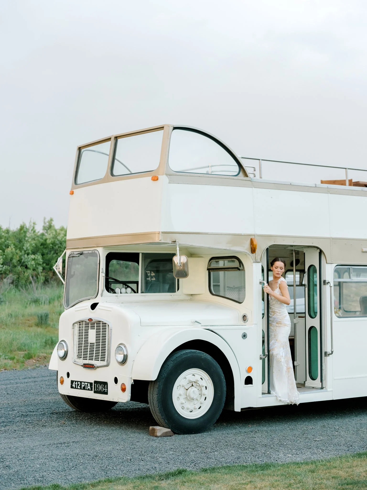 A woman in a white dress standing at the entrance of a vintage white double-decker bus parked on a gravel road with greenery in the background at Greenhouse on Harvard near Spokane, Washington